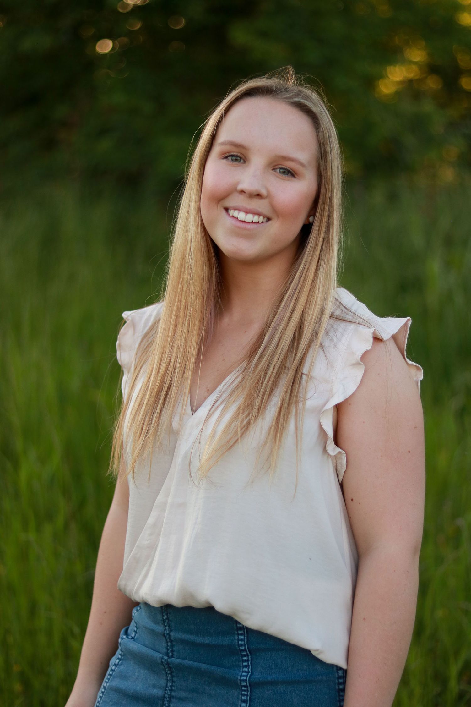 Smiling blonde woman in a white ruffled top standing in a green field at sunset.