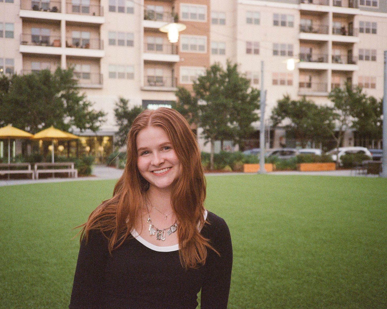 Smiling young woman with red hair standing on green grass in front of a modern apartment building.
