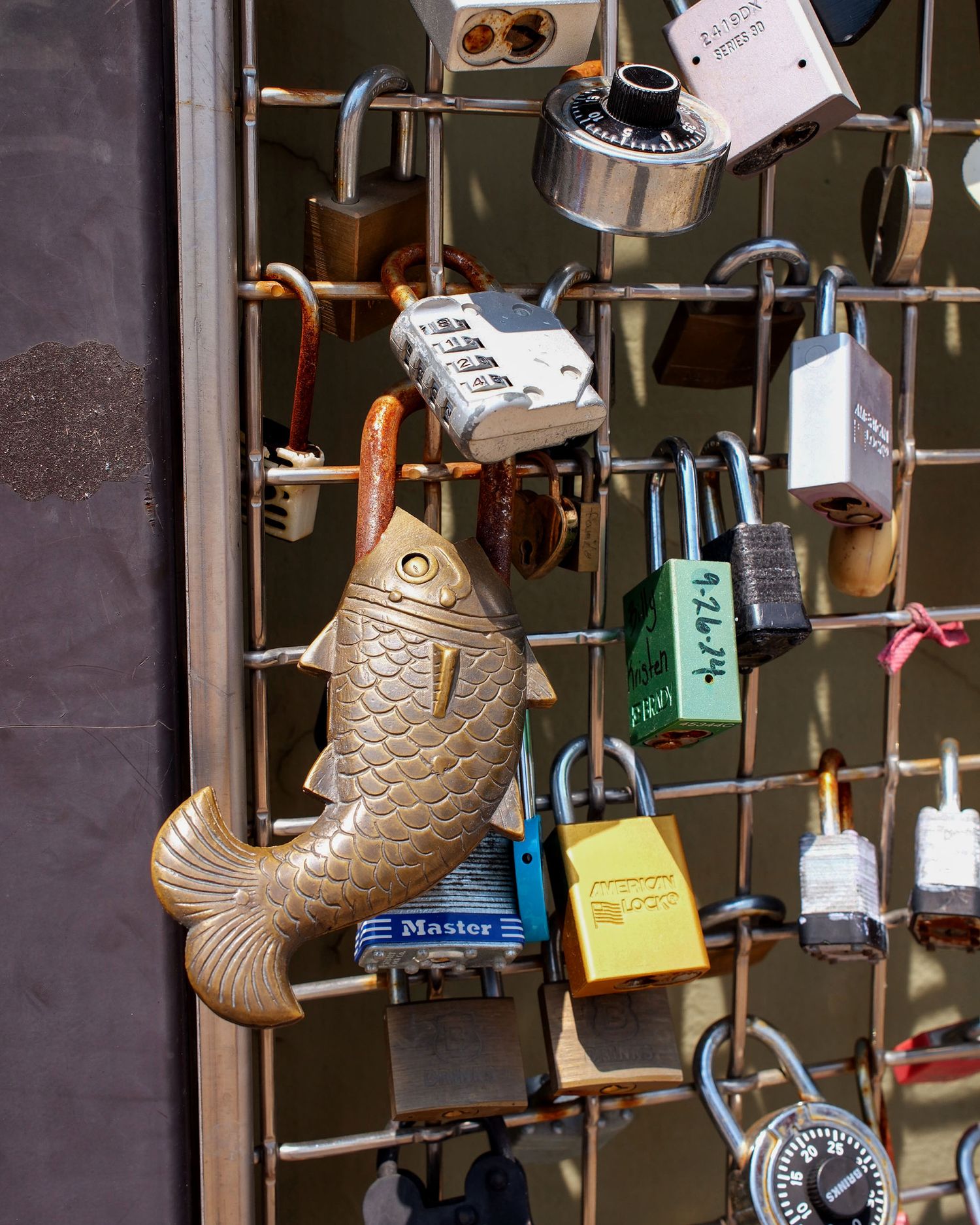 Collection of love locks including a decorative fish padlock on a metal grid fence.