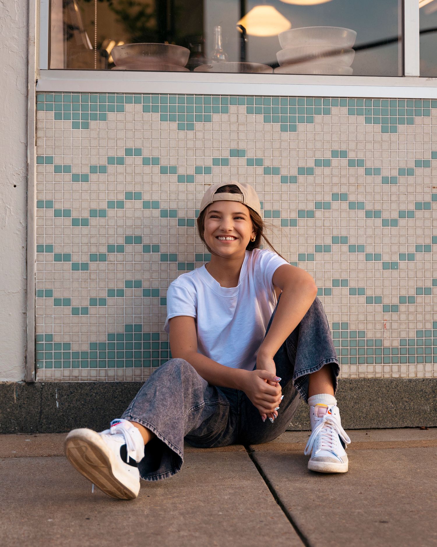 A young person in a white tee, jeans, and snapback sits smiling against a tiled storefront wall.