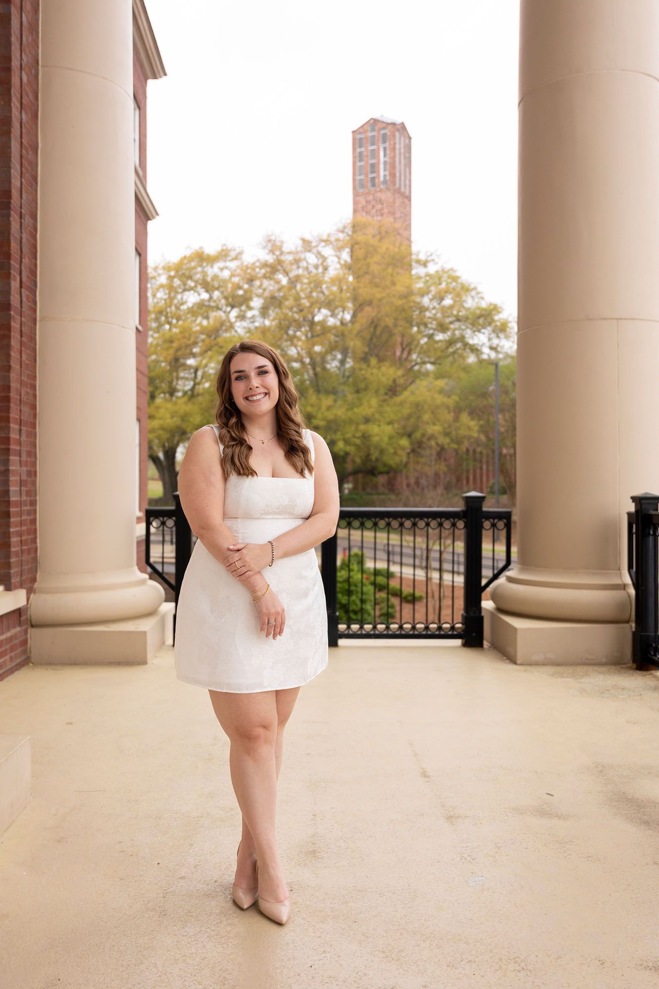 Smiling graduate in white dress standing between columns with a brick bell tower behind her.