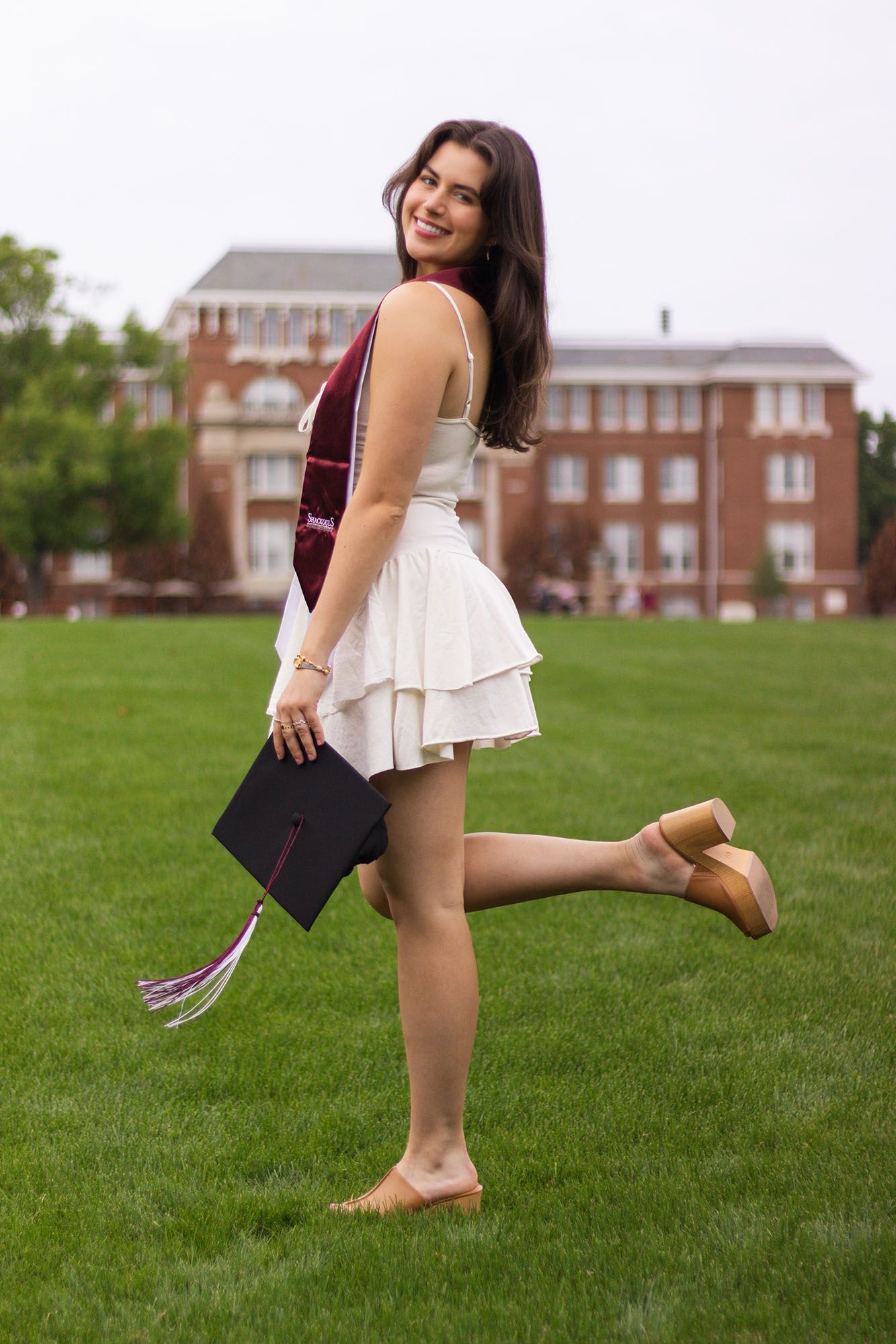 Brunette graduate in white dress holds cap and maroon stole while posing playfully on green lawn.