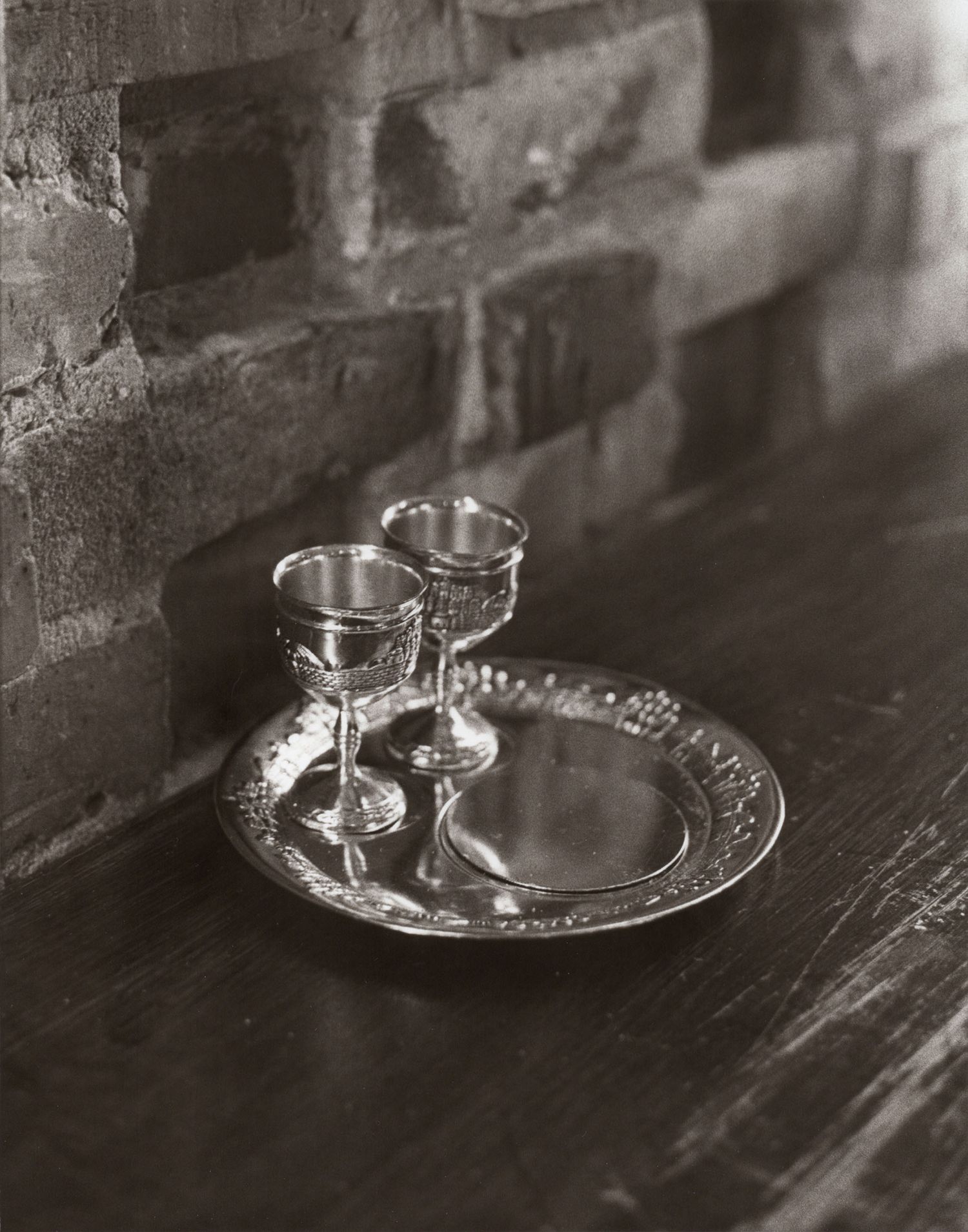 Black and white photo of two ornate silver goblets on a decorative tray beside a brick wall.