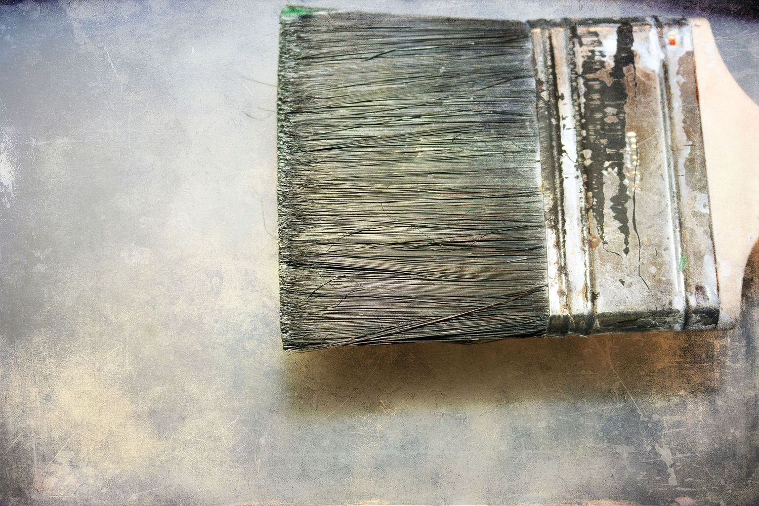 A worn flat paintbrush with dried paint bristles resting on a weathered gray painted surface.