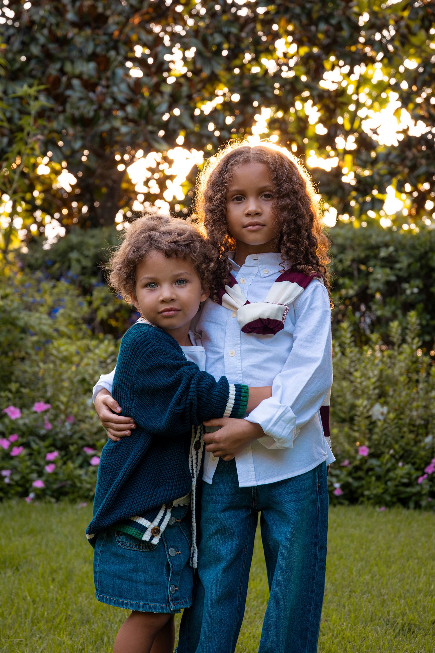Two curly-haired children embrace while posing together in a sunlit garden with flowers.