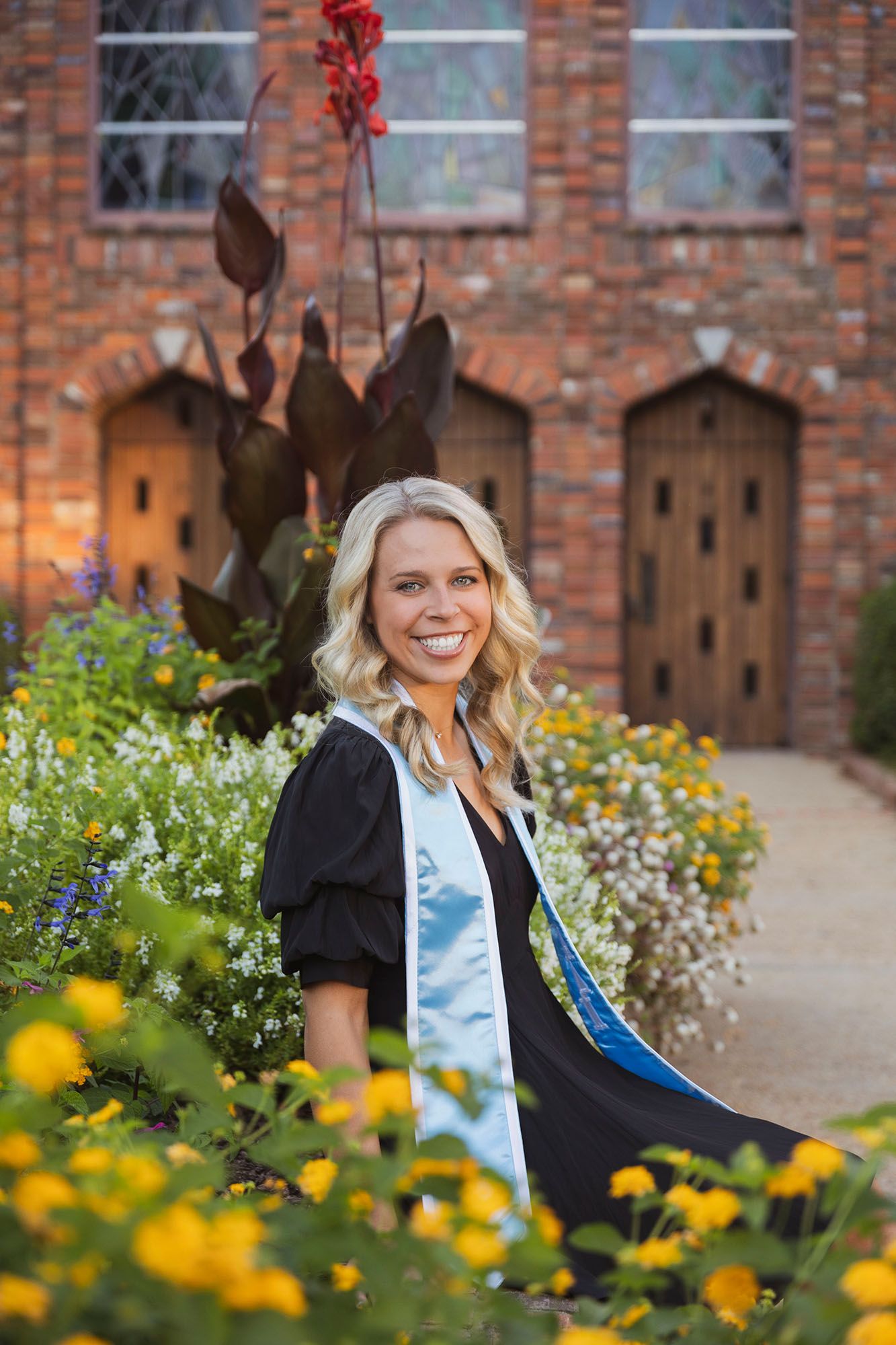 Smiling female graduate in black gown and blue stole poses among colorful flowers at brick building.