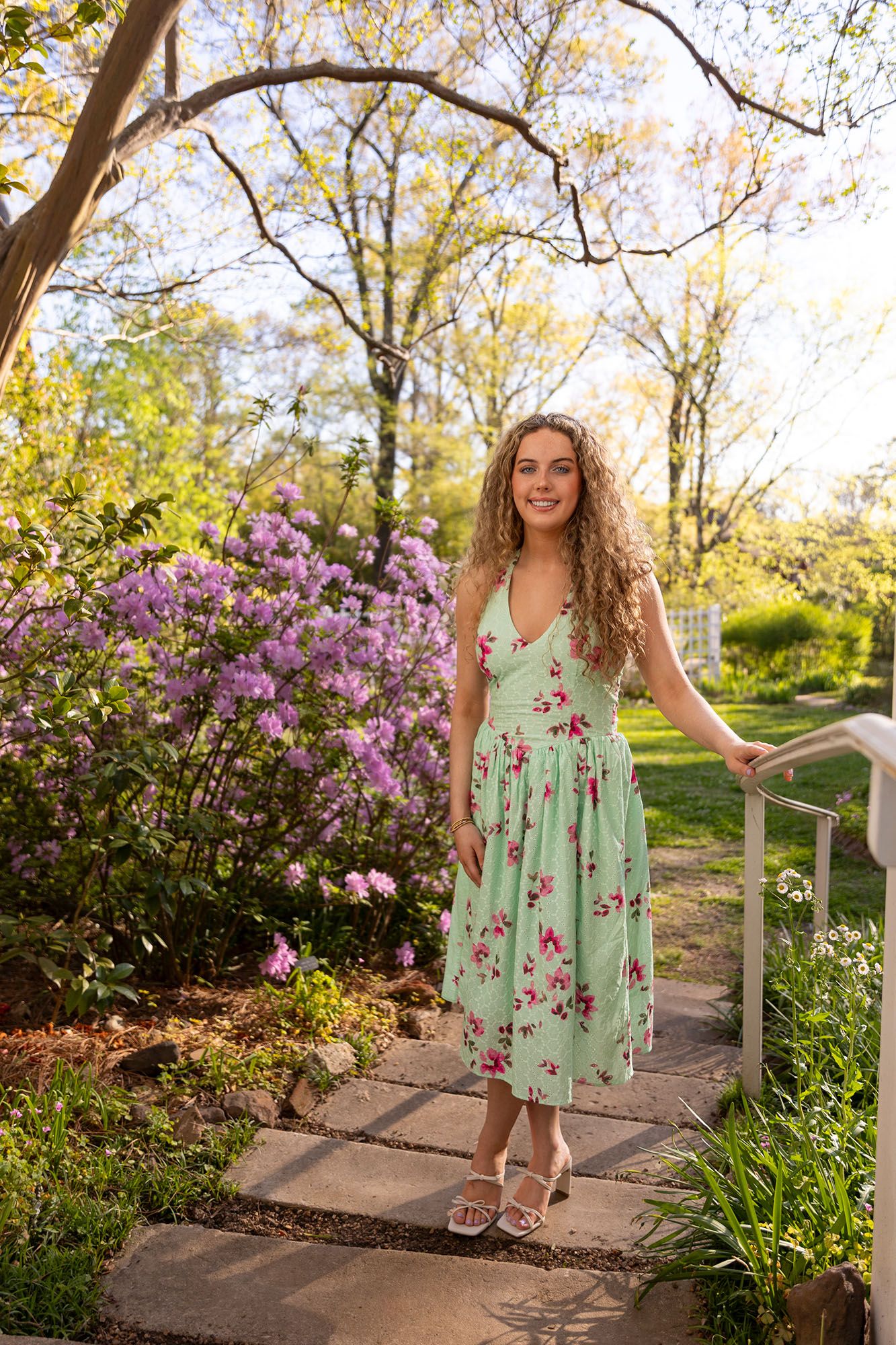 Young woman in mint floral midi dress stands on garden steps surrounded by blooming flowers.