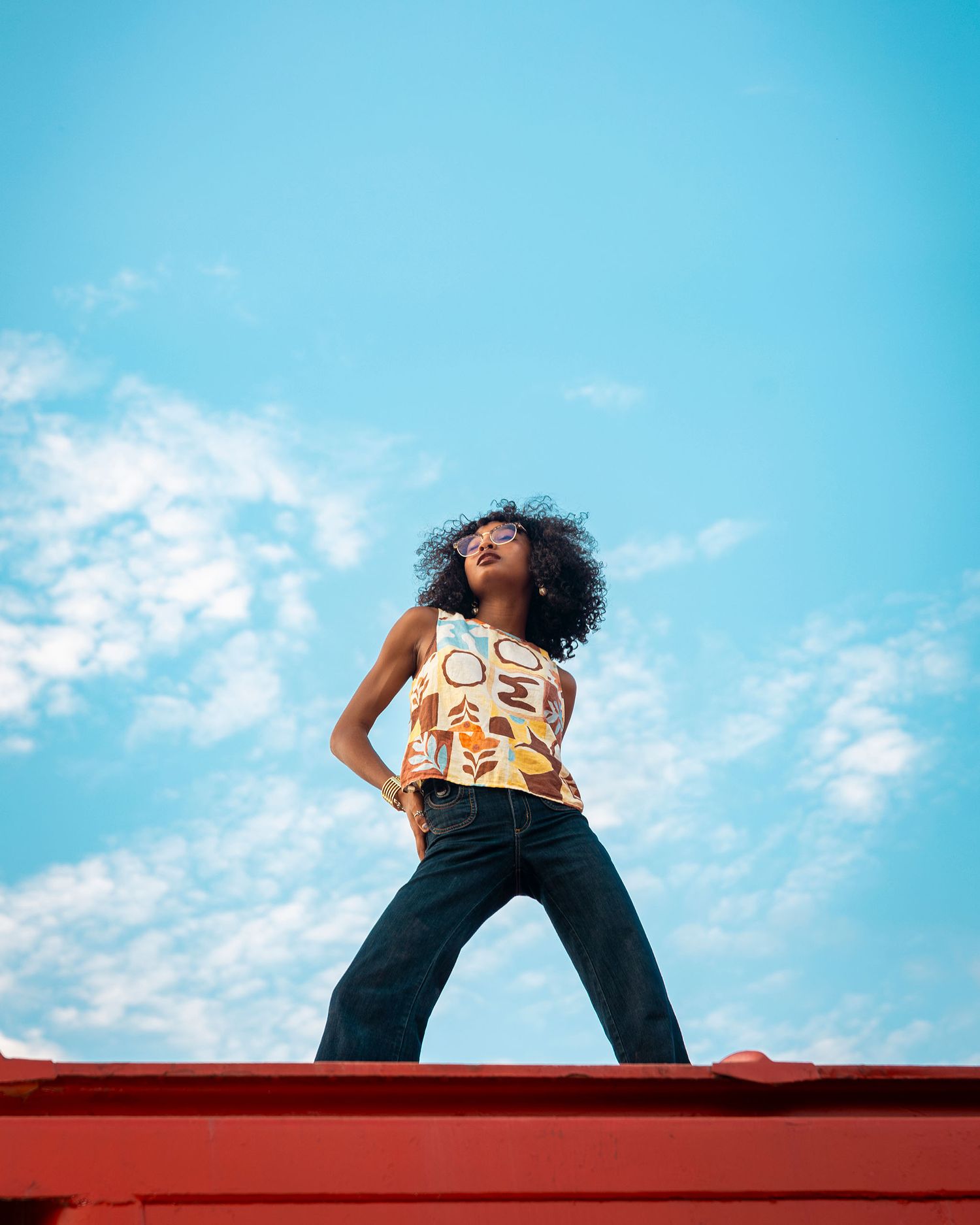 Fashionable woman in geometric print top and wide-leg jeans posing against blue sky on rooftop.