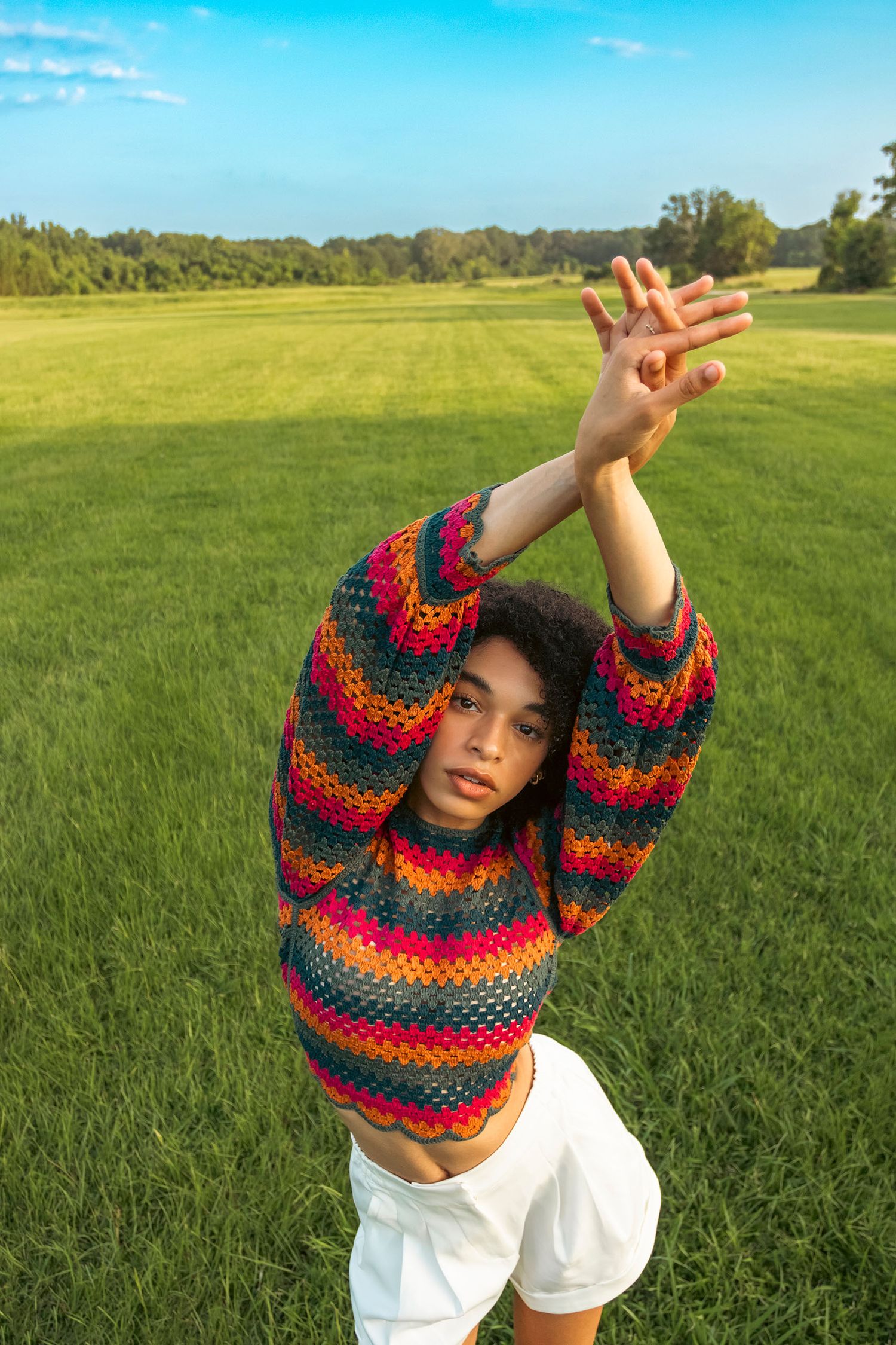 Young woman in colorful striped crochet sweater raising arms overhead in a lush green grass field.