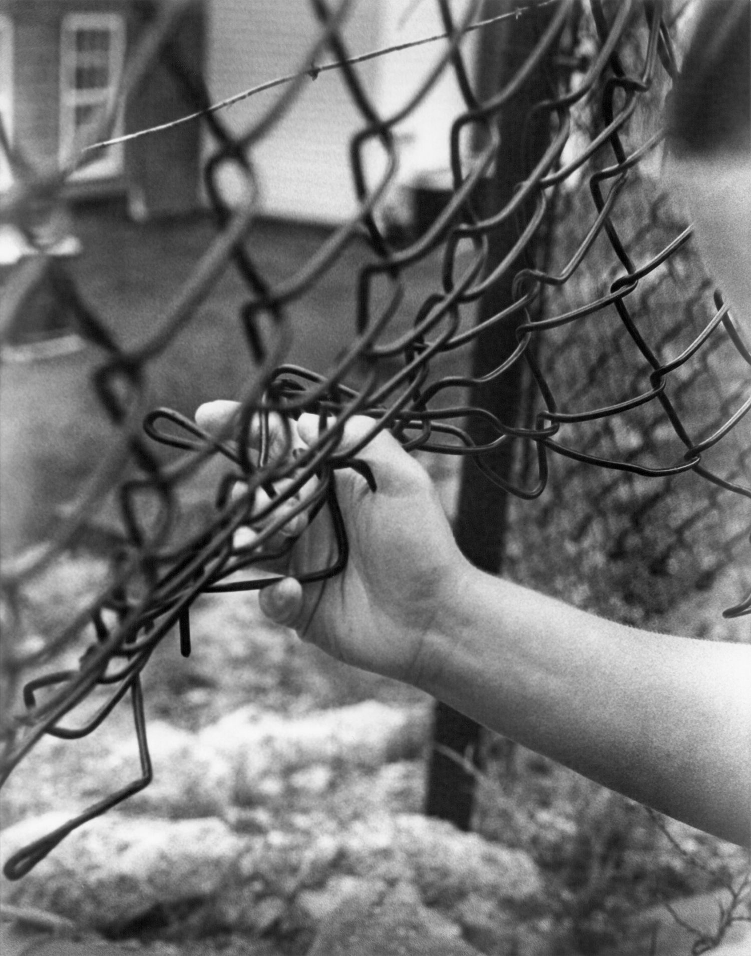 Black and white close-up of a hand gripping a chain-link fence with a house in background.