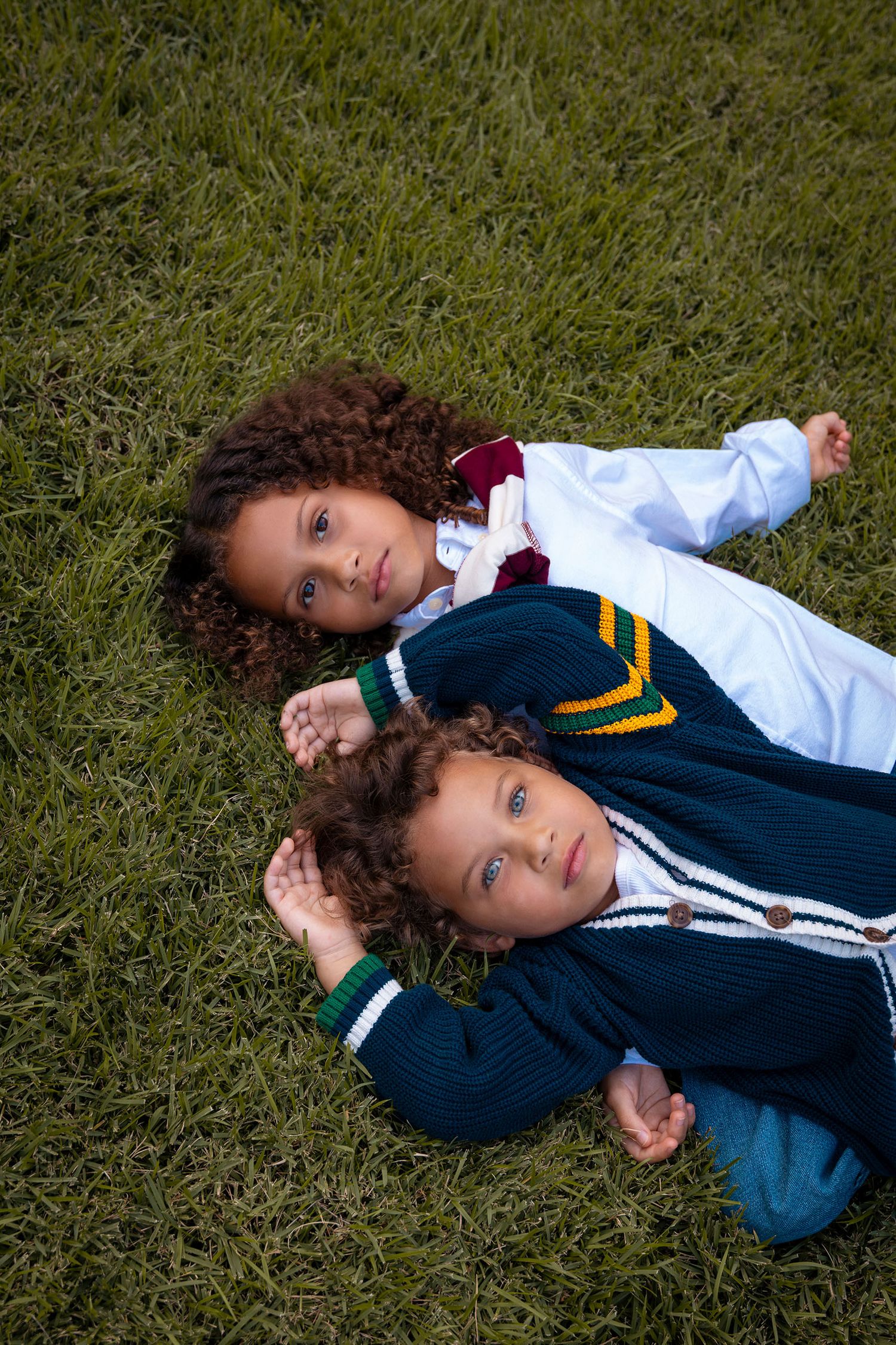 Two curly-haired children with blue eyes lie side by side on green grass outdoors.