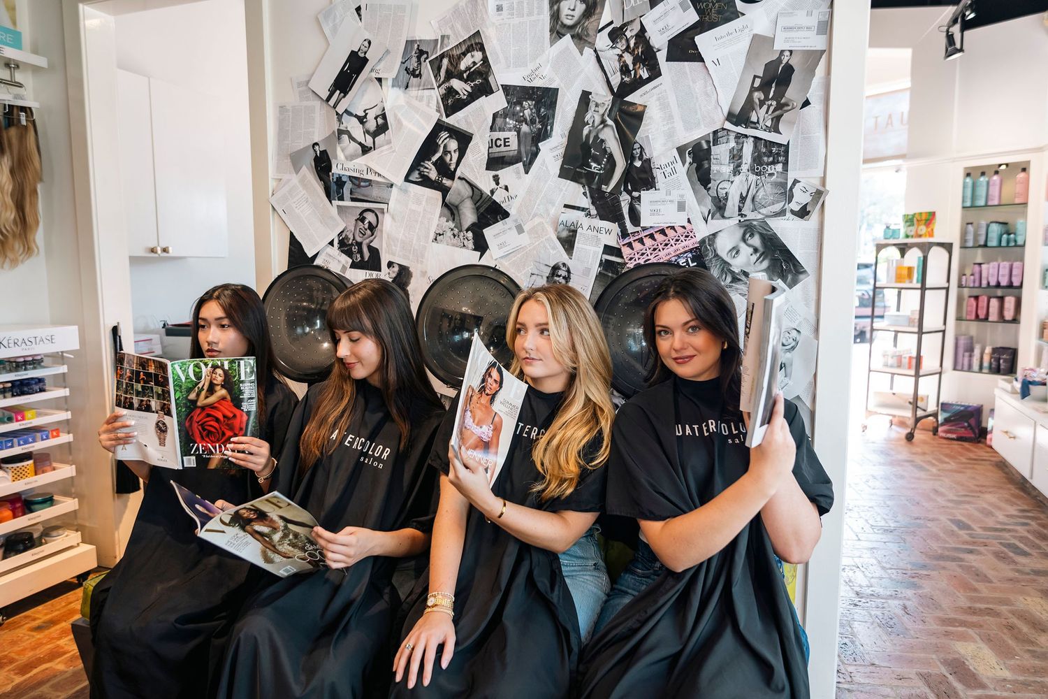 Four women in black salon capes reading magazines in front of a wall covered in black and white photos.