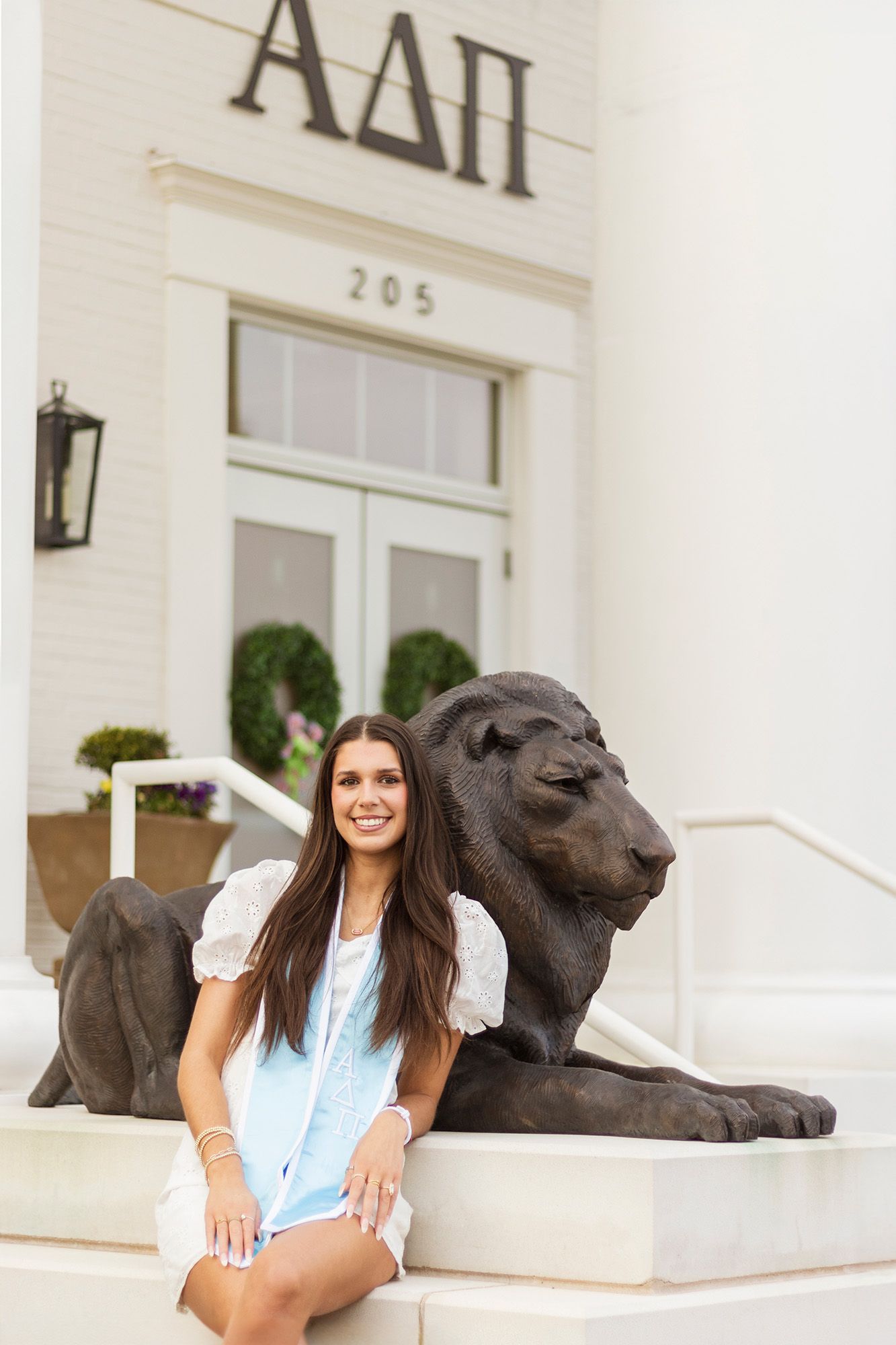Smiling brunette female graduate poses beside lion statue at Alpha Delta Pi sorority house entrance.