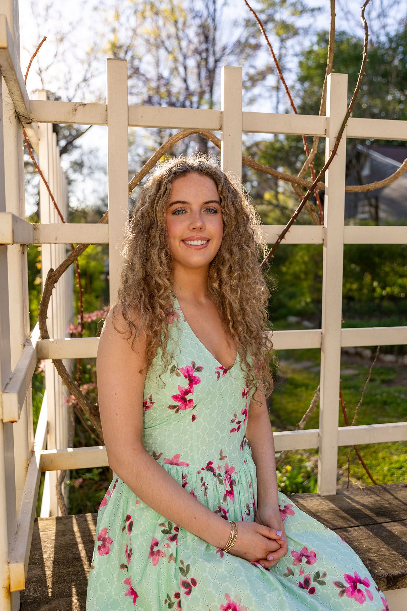 Young woman with curly blonde hair smiles in a mint floral dress seated by a white garden trellis.