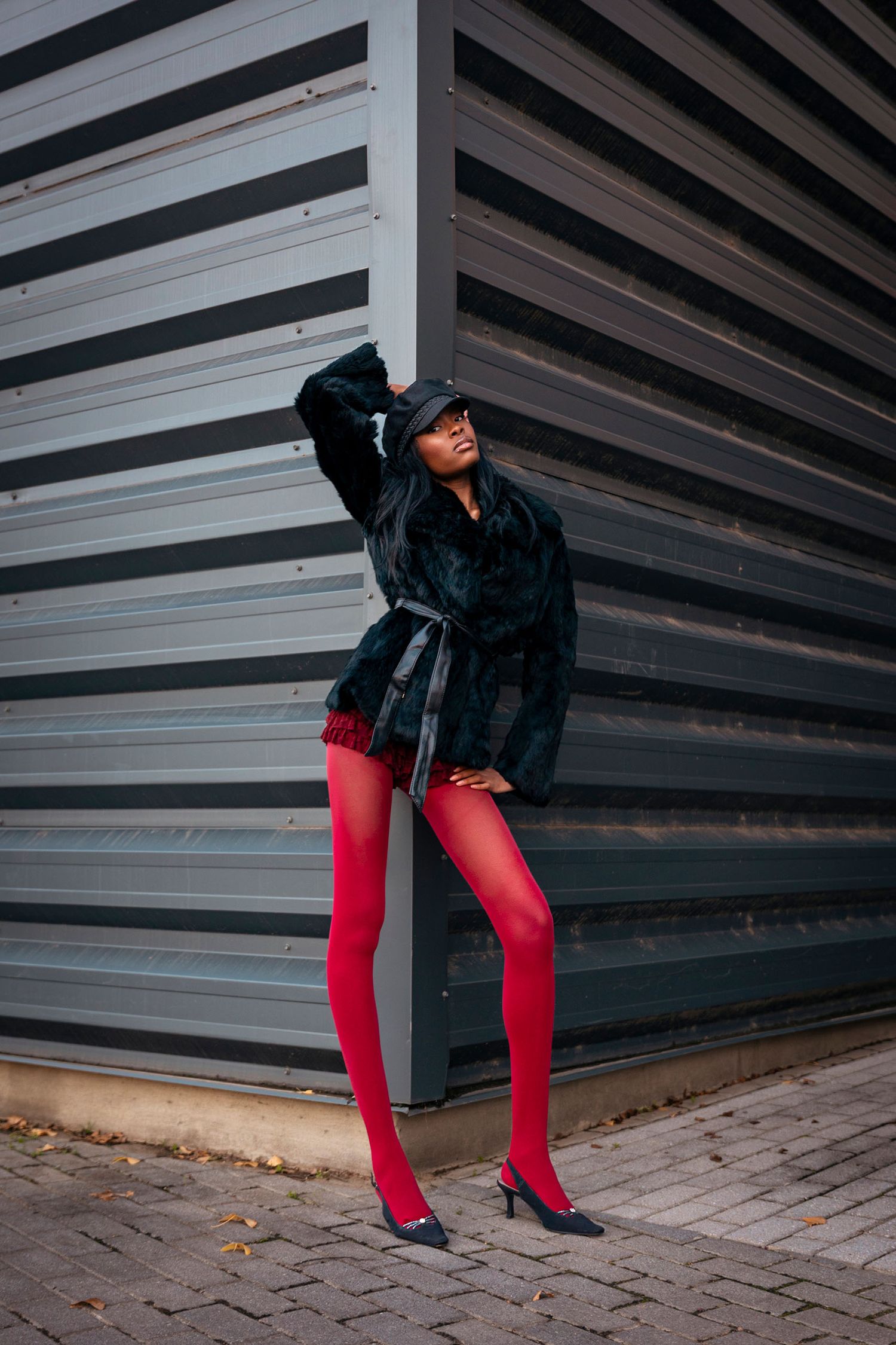 Woman in black fur coat and bold red tights posing against corrugated metal wall wearing black heels and bucket hat.