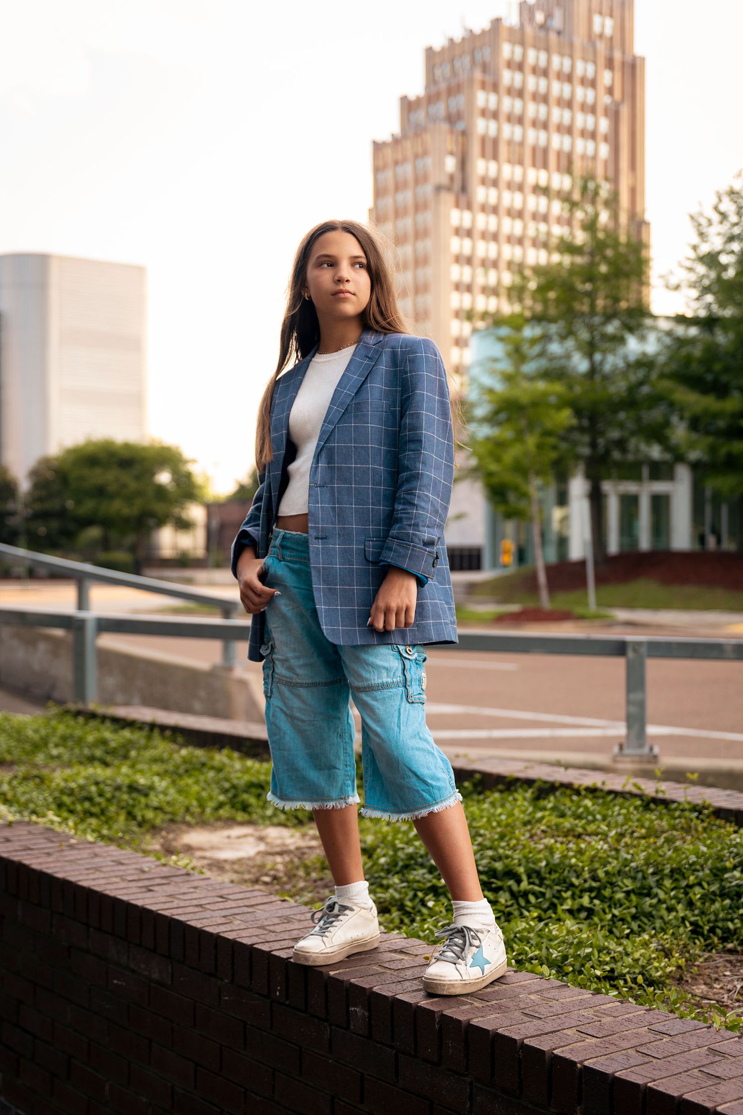 Young woman in blue plaid blazer and denim shorts stands on urban brick ledge.