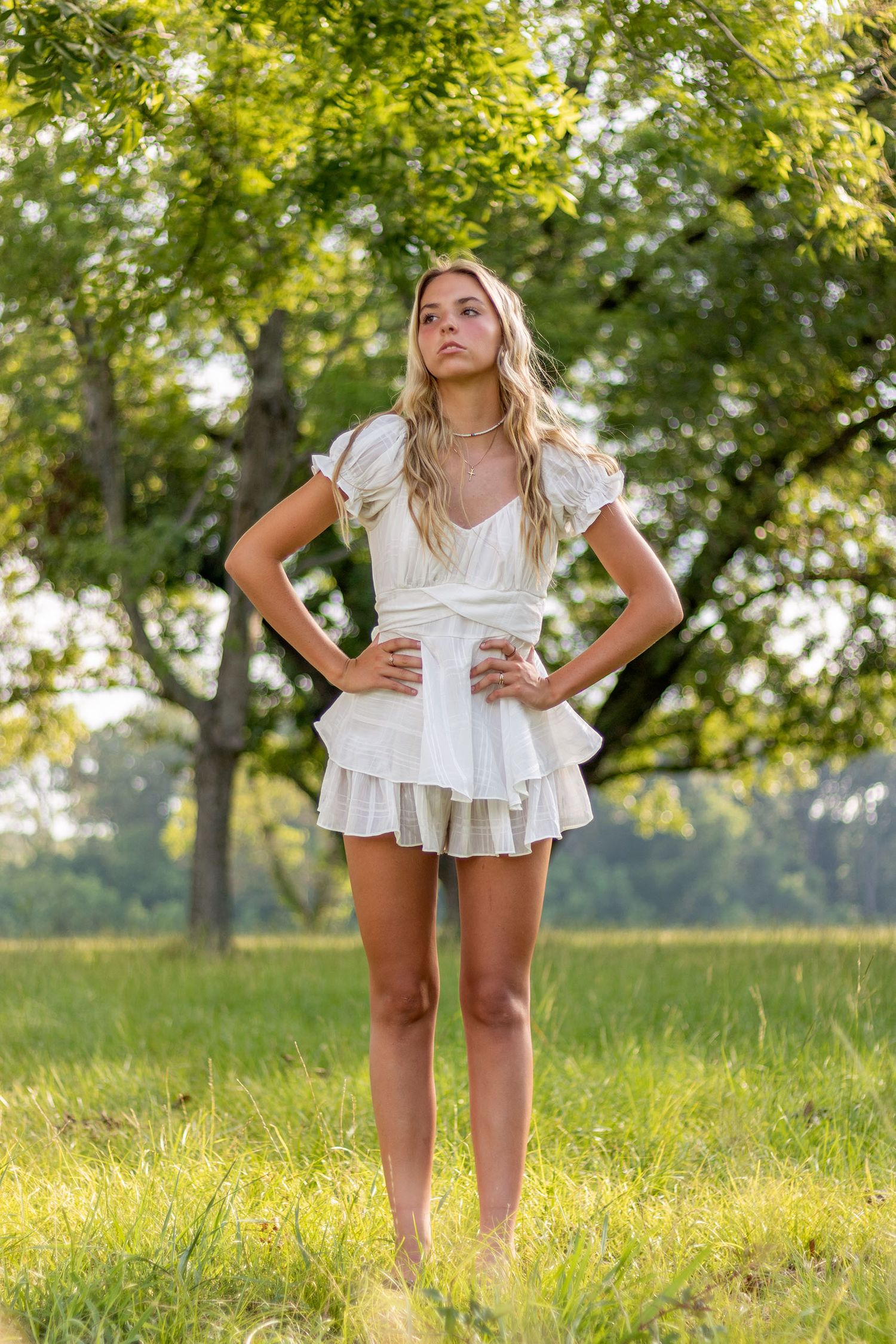 Young blonde woman in white ruffled mini dress posing with hands on hips in a green summer field.
