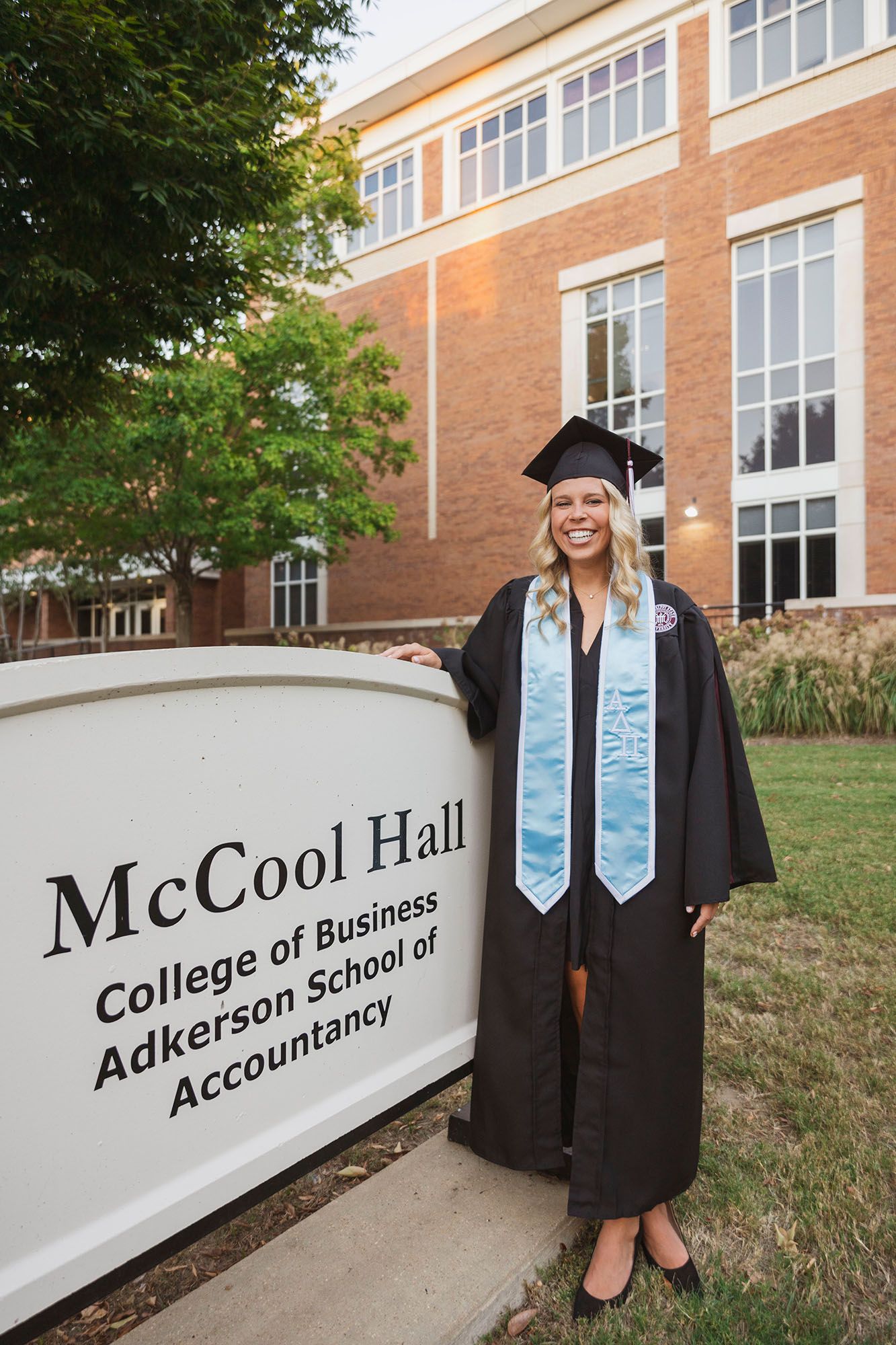 Female graduate in cap and gown poses proudly beside McCool Hall College of Business sign at MSU.