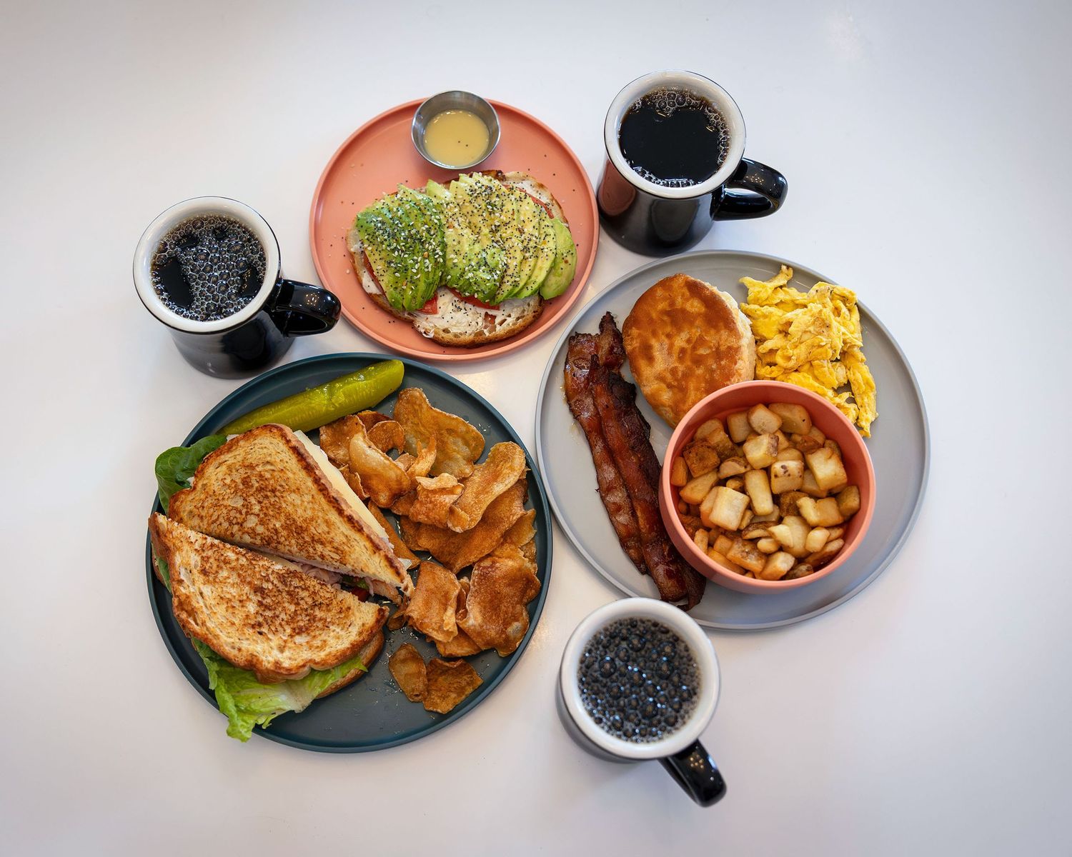 Overhead view of a breakfast spread with sandwiches, eggs, bacon, avocado toast, and black coffee.