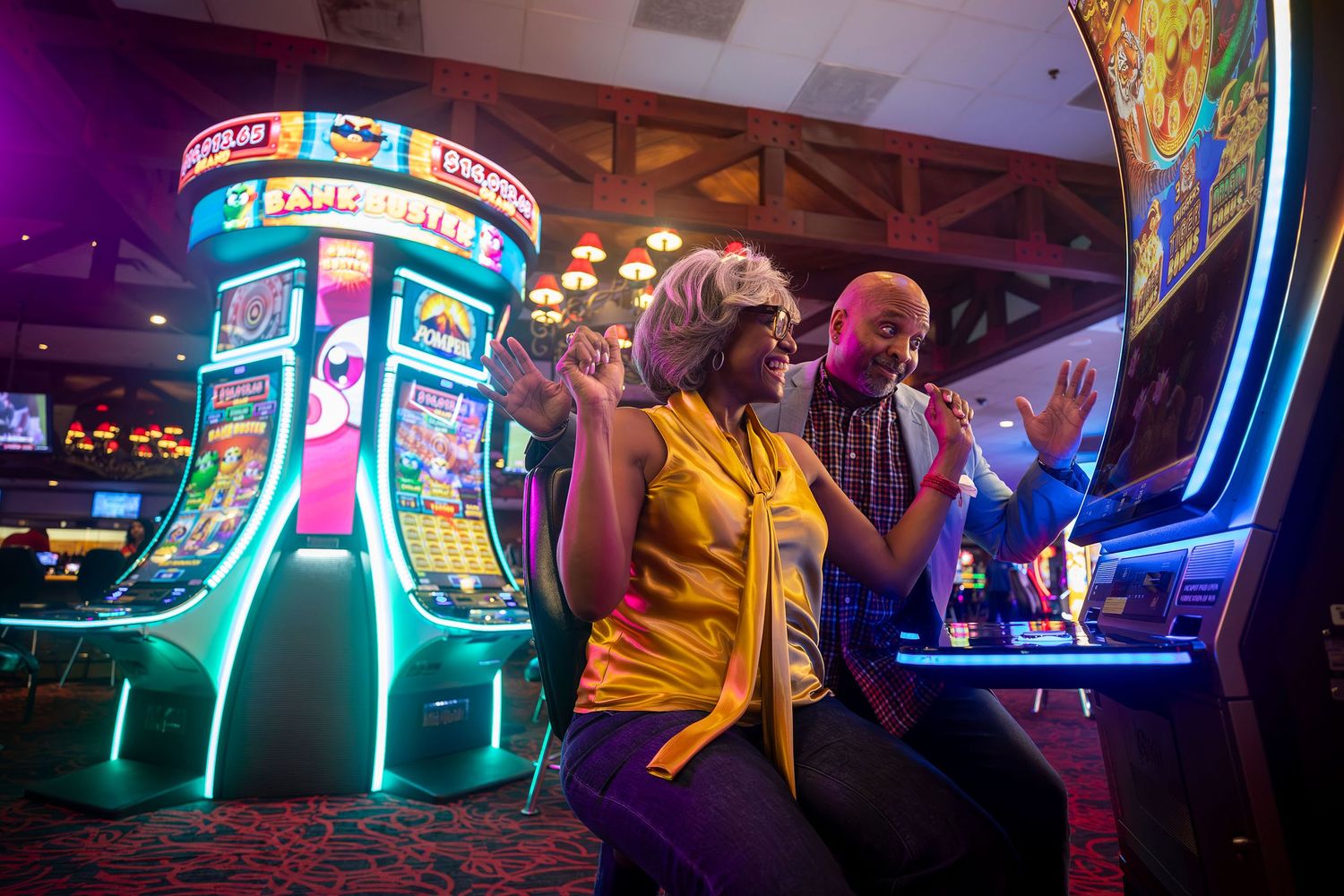 Two excited guests celebrate at colorful slot machines inside a vibrant casino floor.