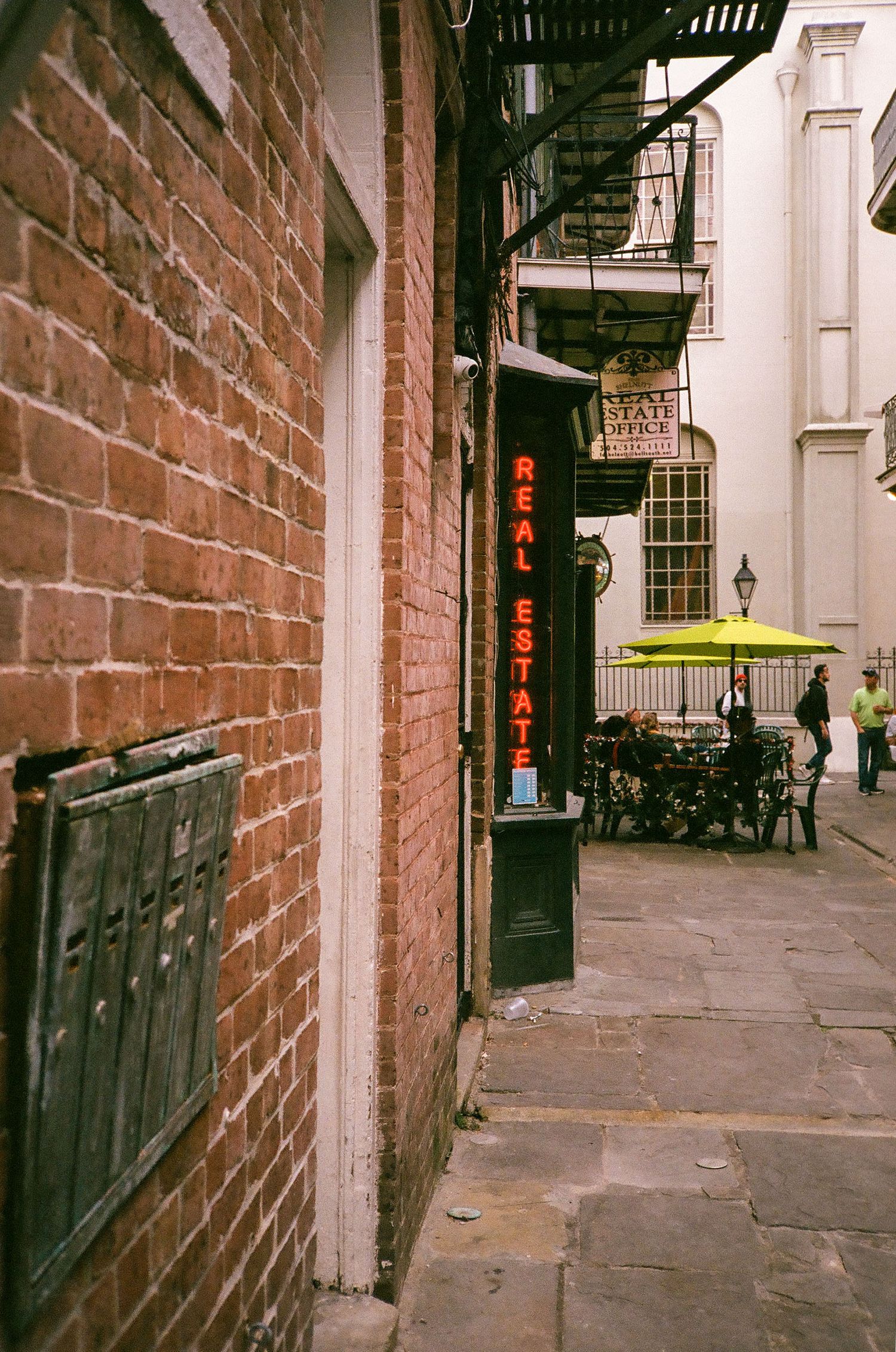 A narrow brick alley in New Orleans with a red neon Real Estate sign and yellow umbrella cafe.