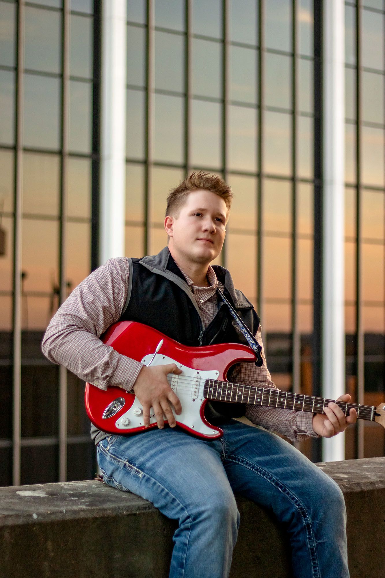 Young man sitting on ledge holding red electric guitar at sunset with glass building backdrop.