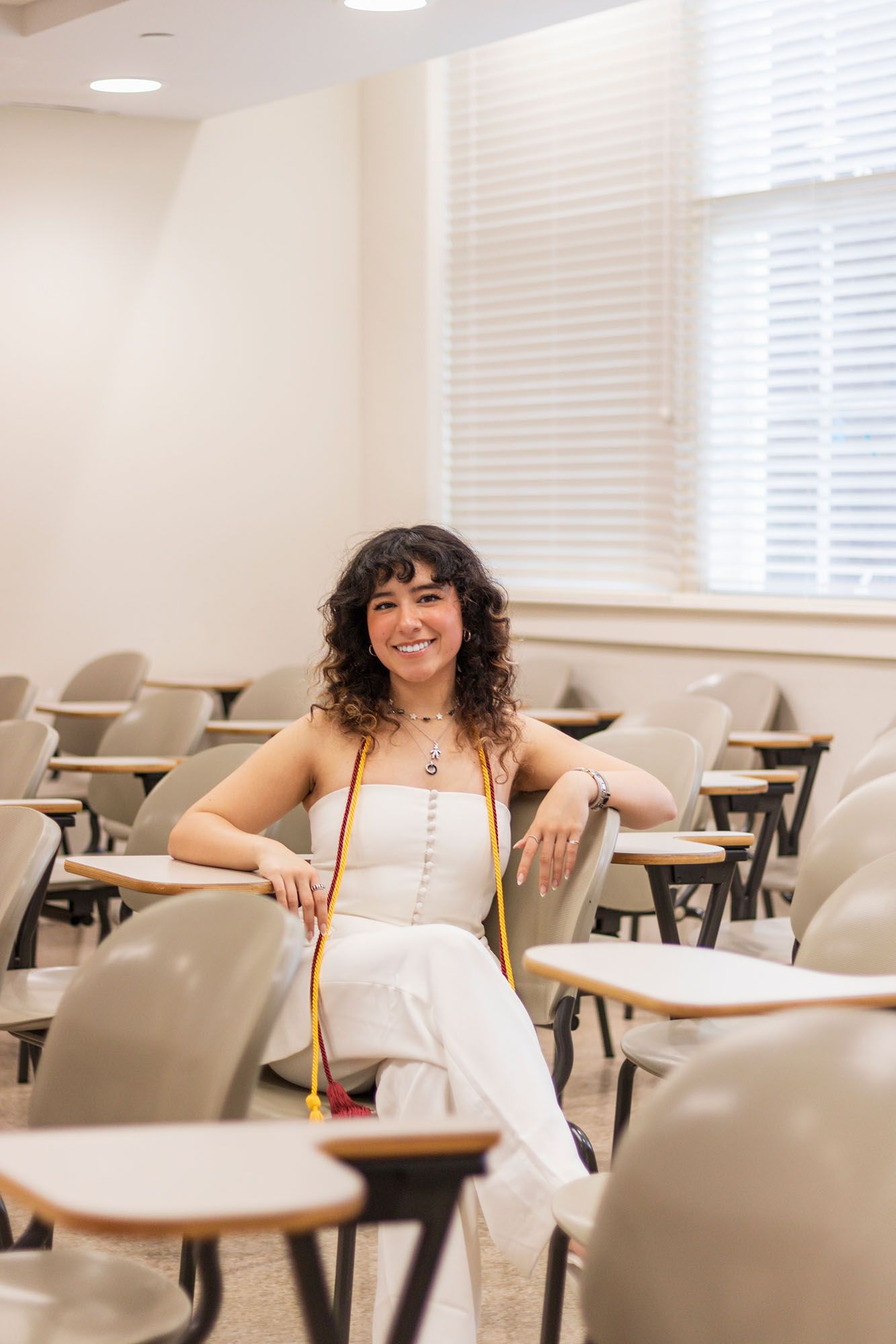 Smiling female graduate with honor cords sits relaxed in empty university classroom.
