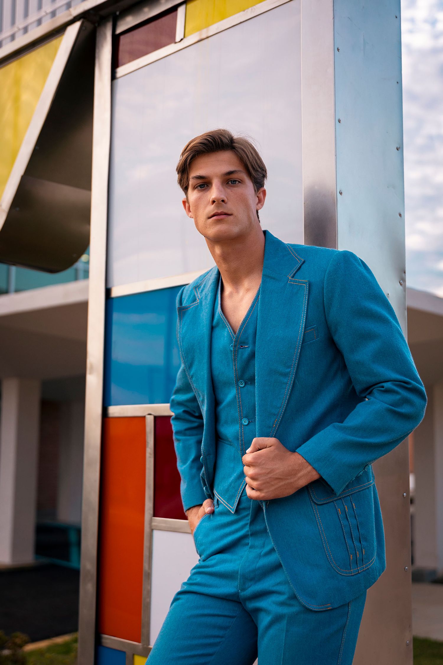 Male model in teal three-piece suit poses against colorful geometric building exterior.