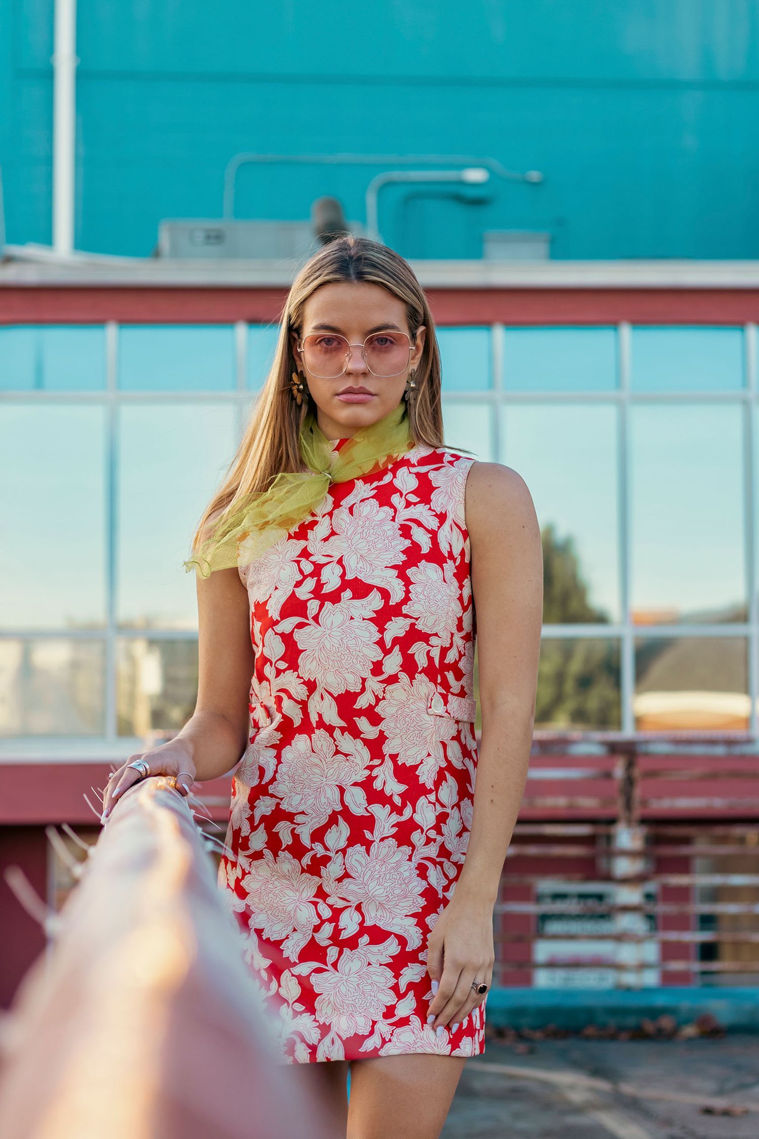 A woman in a red floral dress and yellow scarf poses outdoors near a teal building.