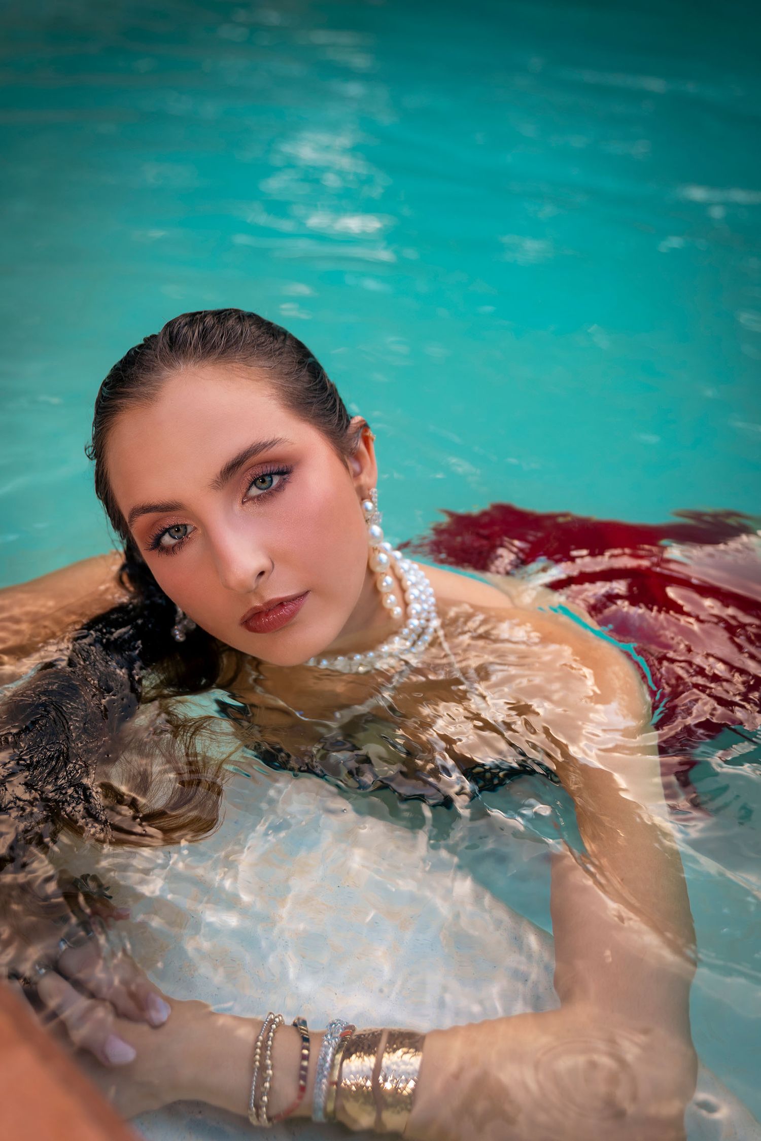 Woman in red dress and pearl necklace posing in turquoise swimming pool with wet hair and gold bracelets.