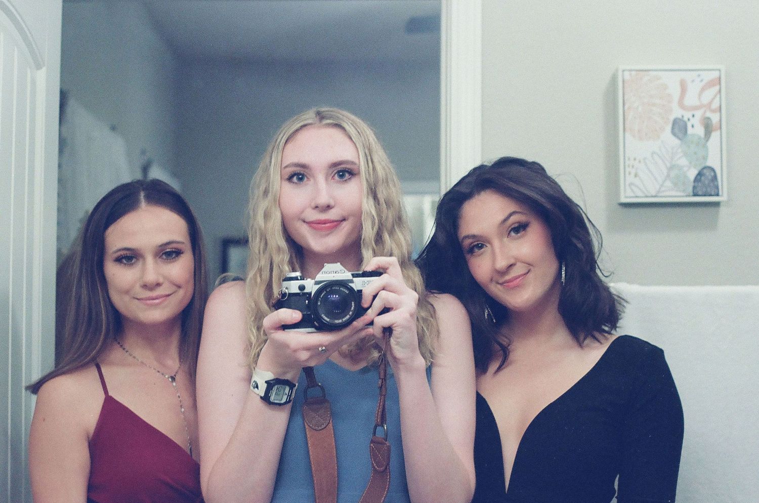 Three young women smiling together, one holding a film camera, taking a mirror selfie.