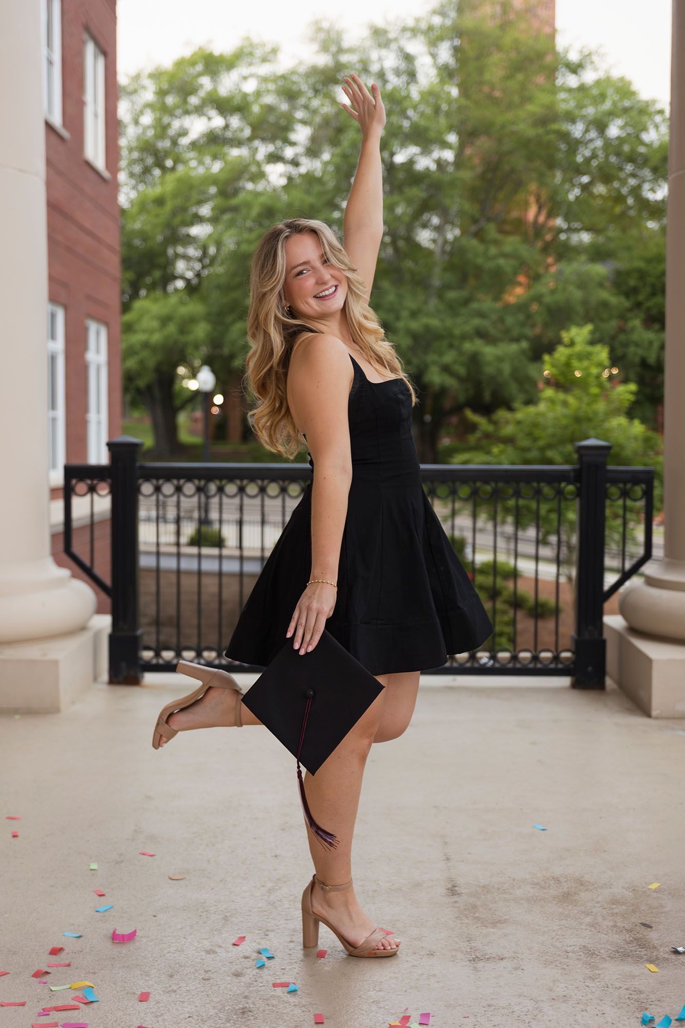 Joyful female graduate in black dress celebrates with cap amid confetti on campus.