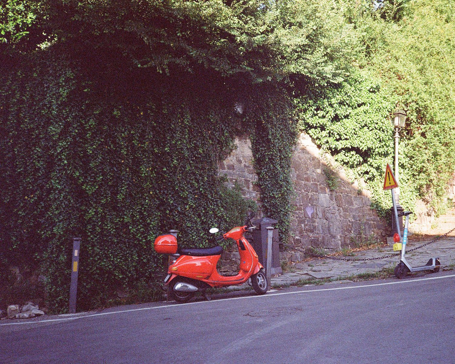 A red Vespa scooter parked beside an ivy-covered stone wall along a quiet street.