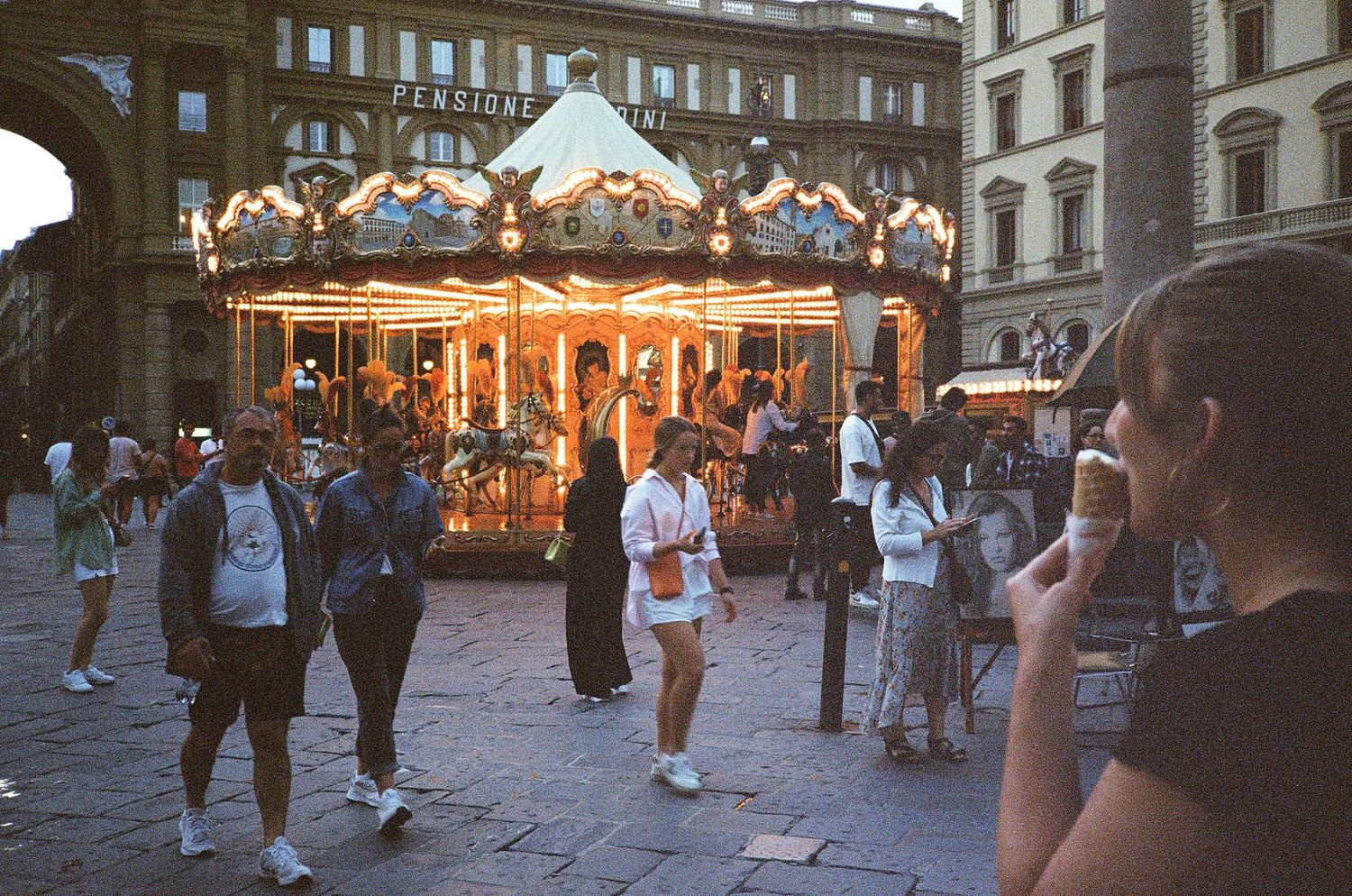 Tourists gathered around a lit carousel at dusk in a historic European city square.