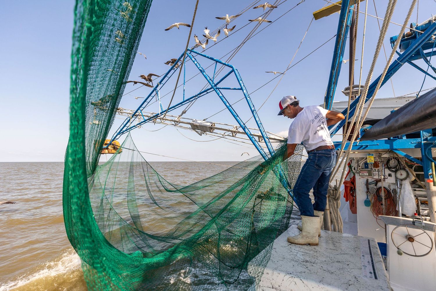 A fisherman hauls a large green net on a shrimp boat on calm open waters under a clear blue sky.