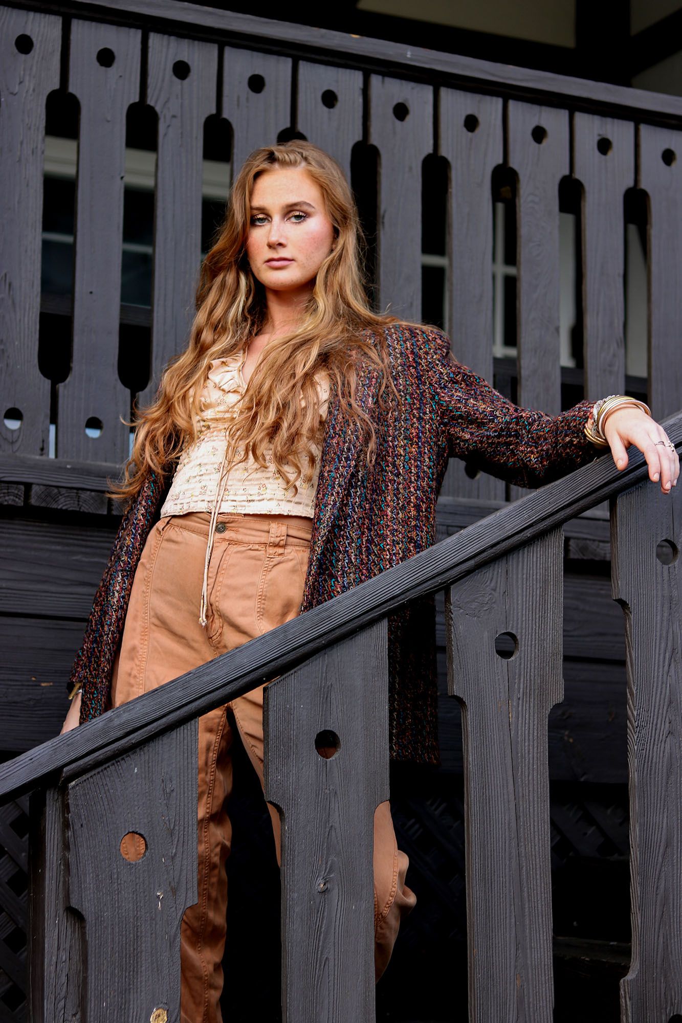 Woman with long wavy hair in tweed cardigan and tan suede skirt leans on dark wooden stair railing.