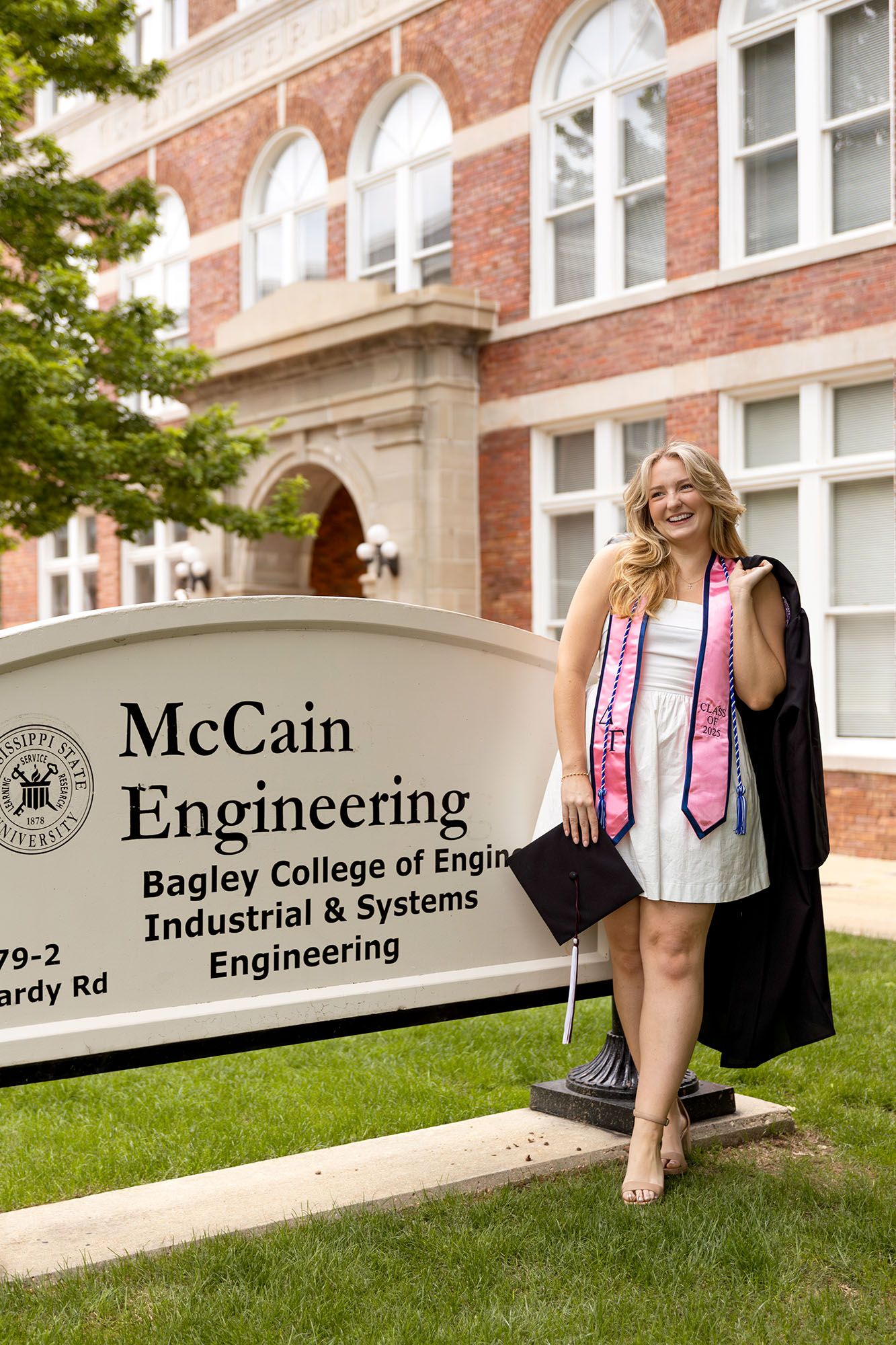 Female graduate poses by McCain Engineering Bagley College sign in black gown.