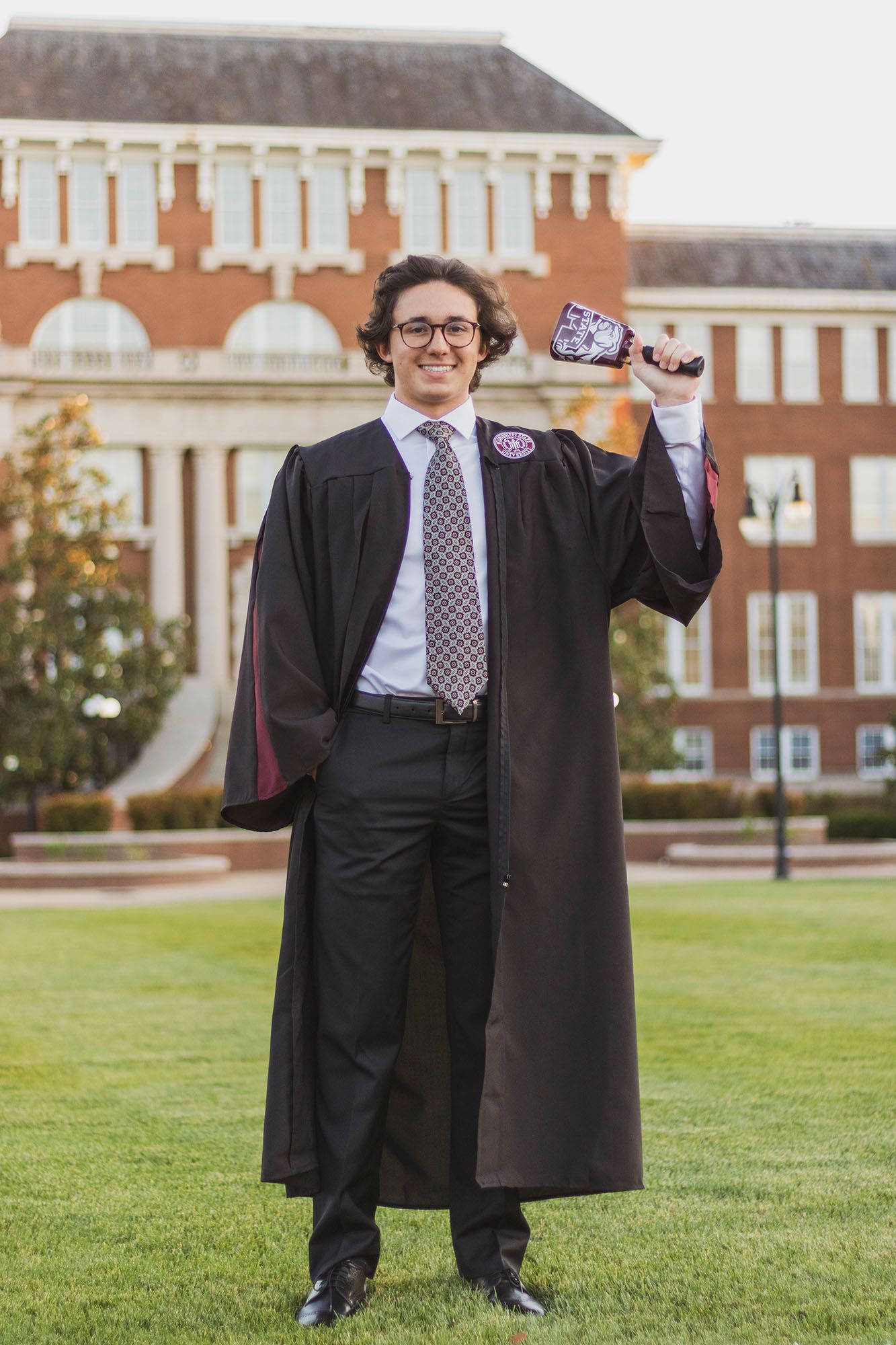 Smiling graduate in black gown holding diploma on green lawn with red brick university building behind him.
