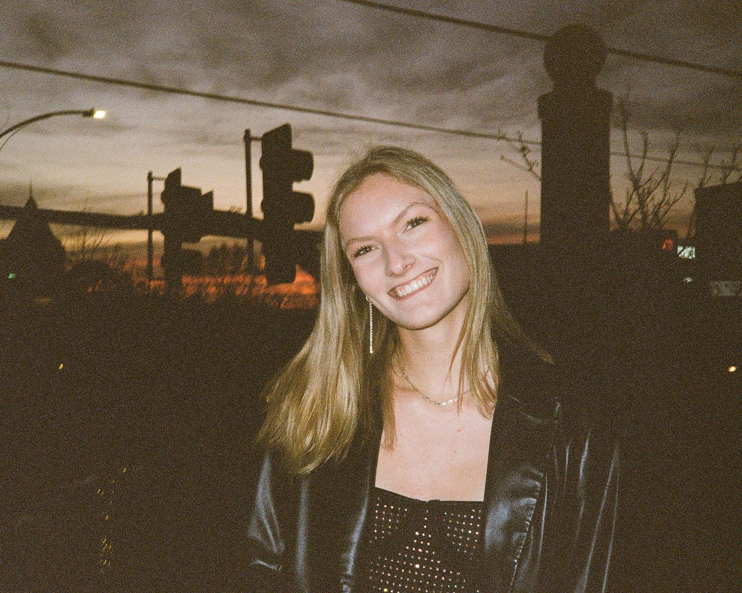 Smiling young woman in a leather jacket standing outdoors at dusk with a colorful sunset sky.