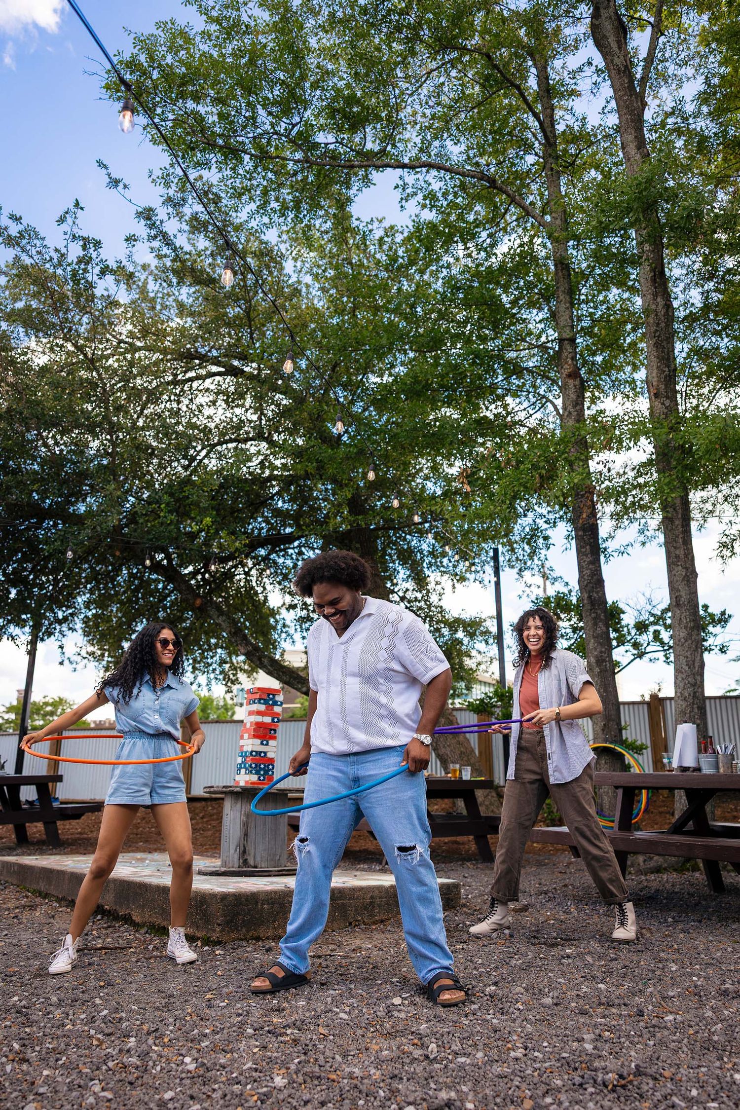 Three friends laughing and hula hooping outdoors at a casual restaurant patio under tall trees.