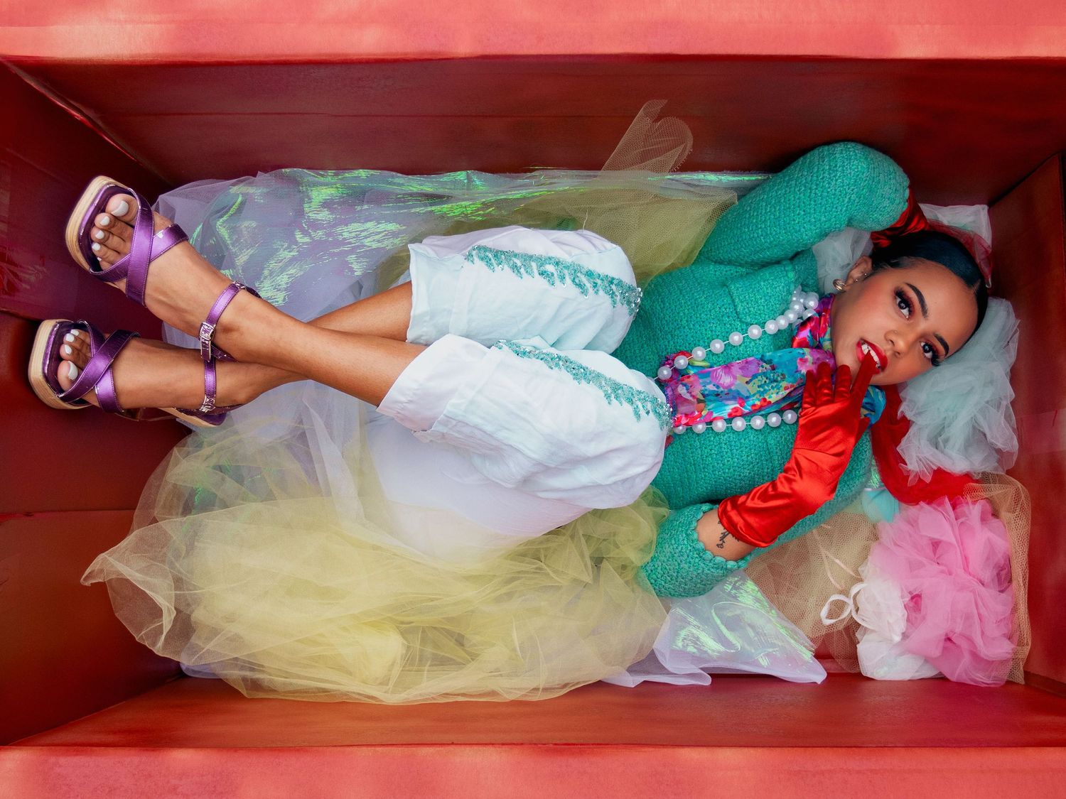 Woman in colorful tulle skirt and teal jacket poses lying in pink box with vibrant accessories.