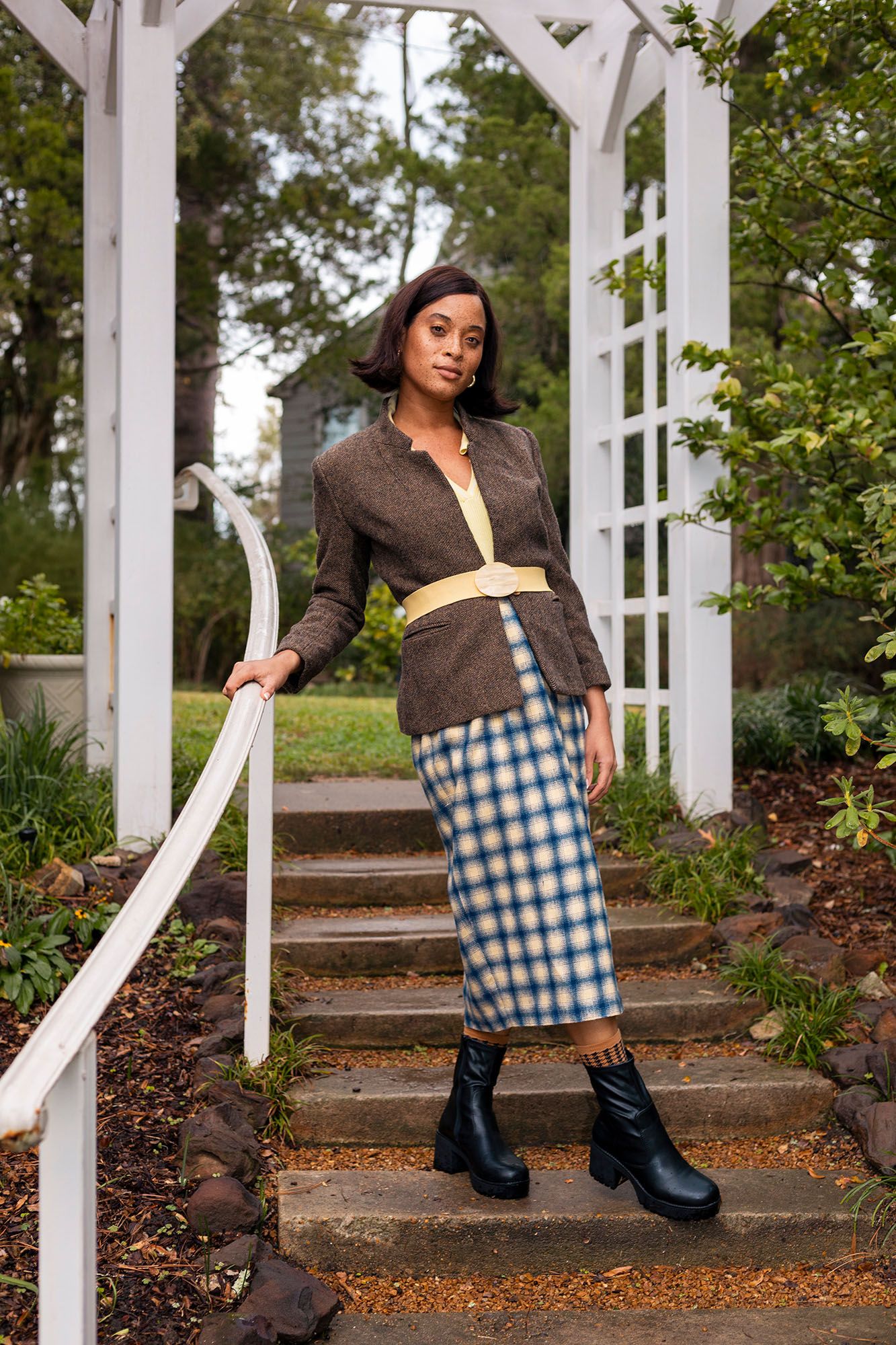 Woman in brown blazer and plaid midi skirt stands on garden steps with black boots.