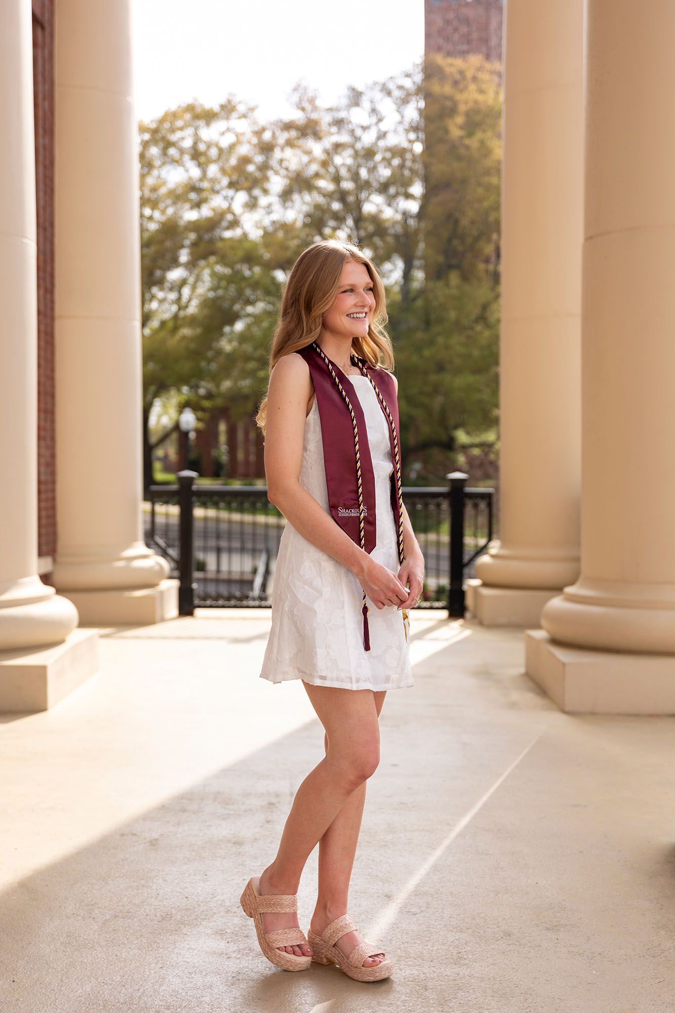 Joyful female graduate in white dress and maroon stole stands between campus columns.