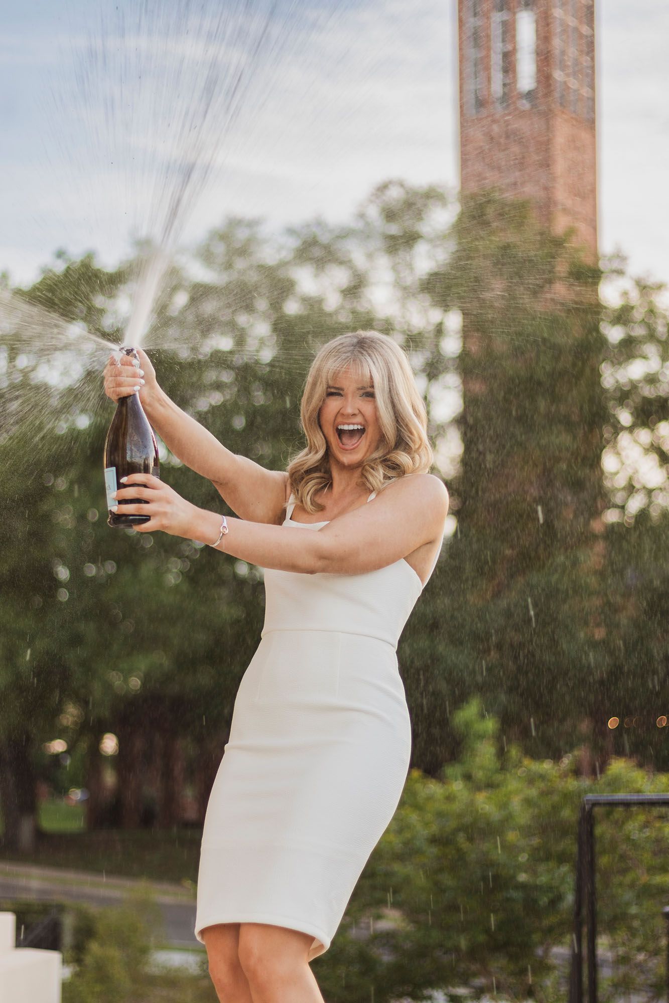 Woman in white dress joyfully popping champagne bottle outdoors with trees and brick tower in background.