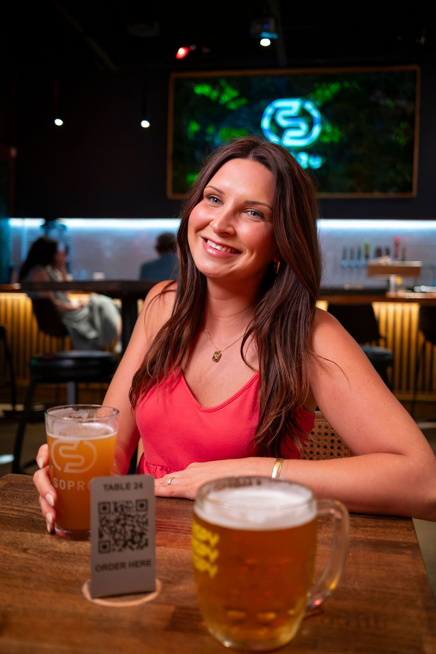 A smiling woman in a pink top holding a beer at a modern bar with QR code table ordering.
