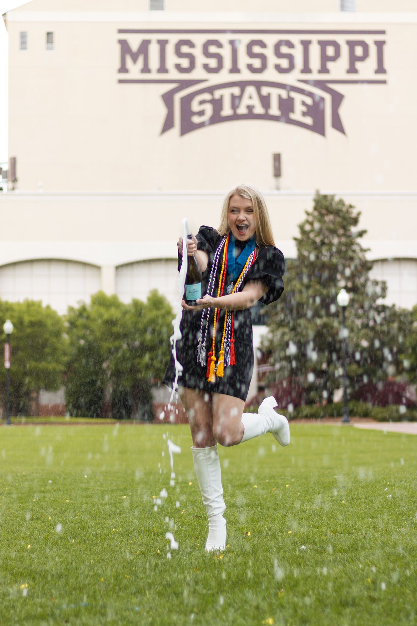 Excited graduate in cap and gown celebrates with champagne spray in front of Mississippi State sign.