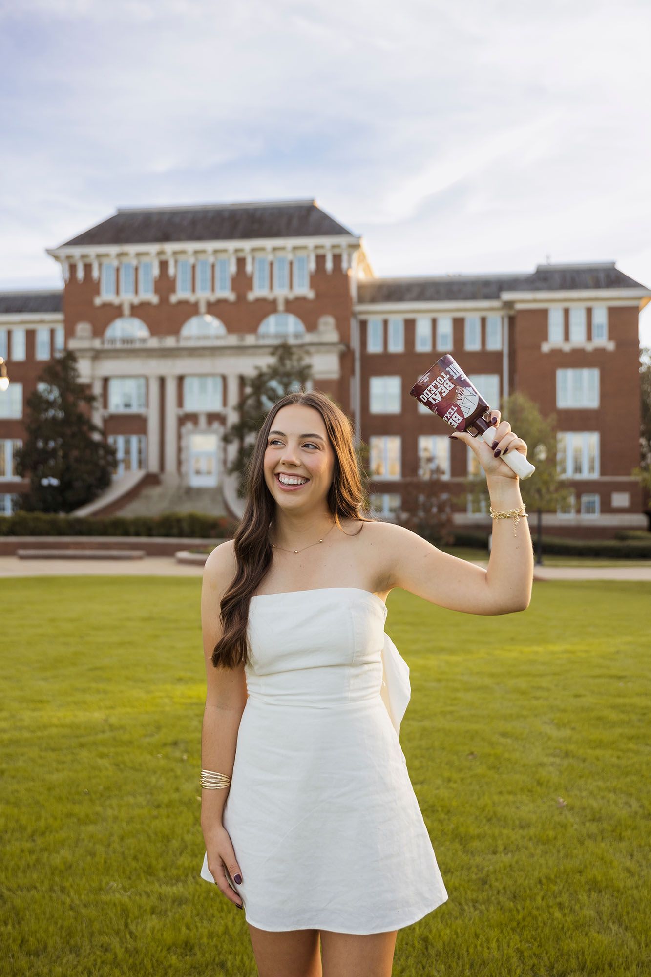 Smiling graduate in white dress holding a cup on a green campus lawn at sunset.