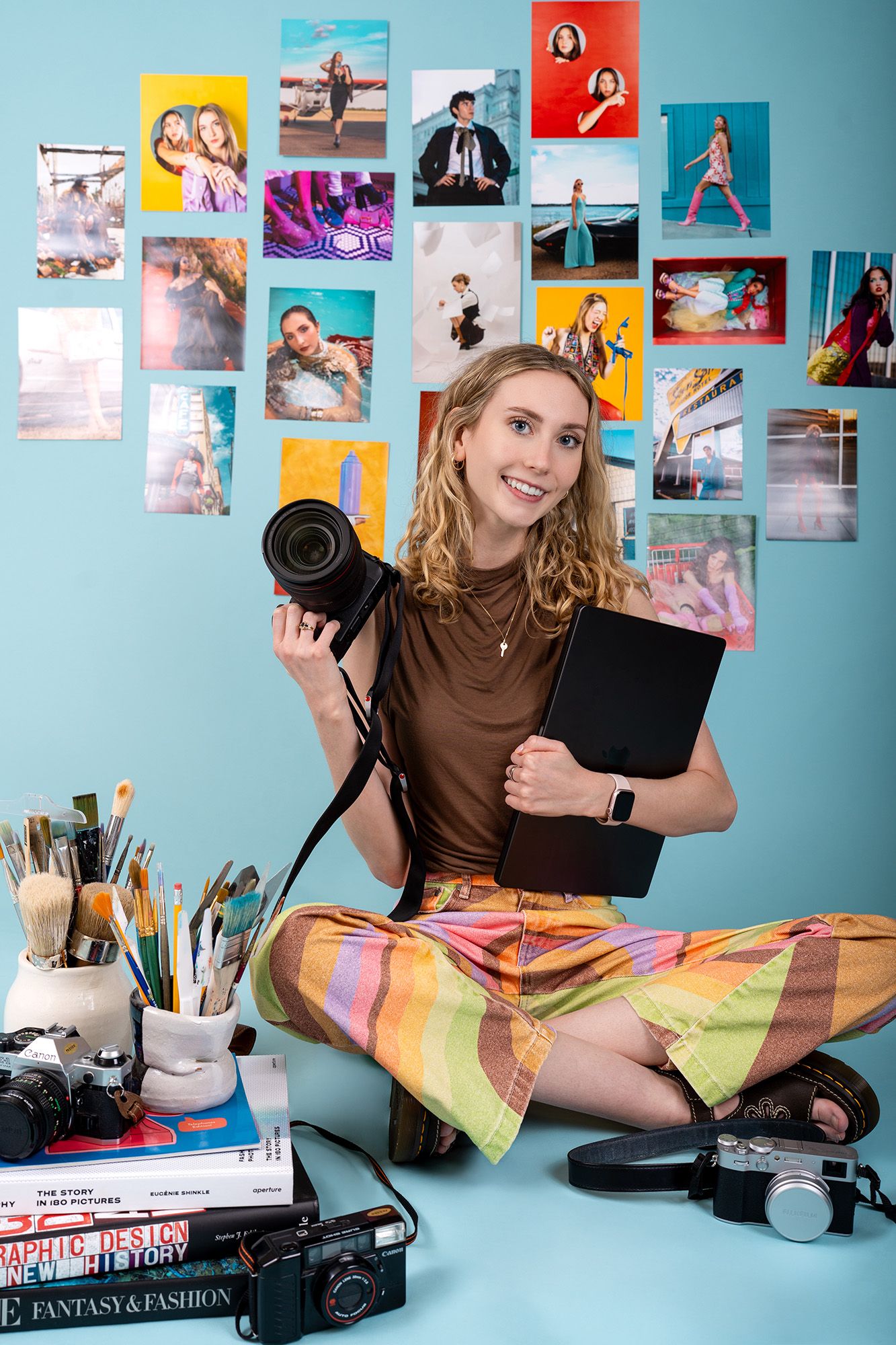 Michelle Daschbach, a photographer sitting cross-legged holding a camera and laptop, surrounded by colorful photos on a blue wall.