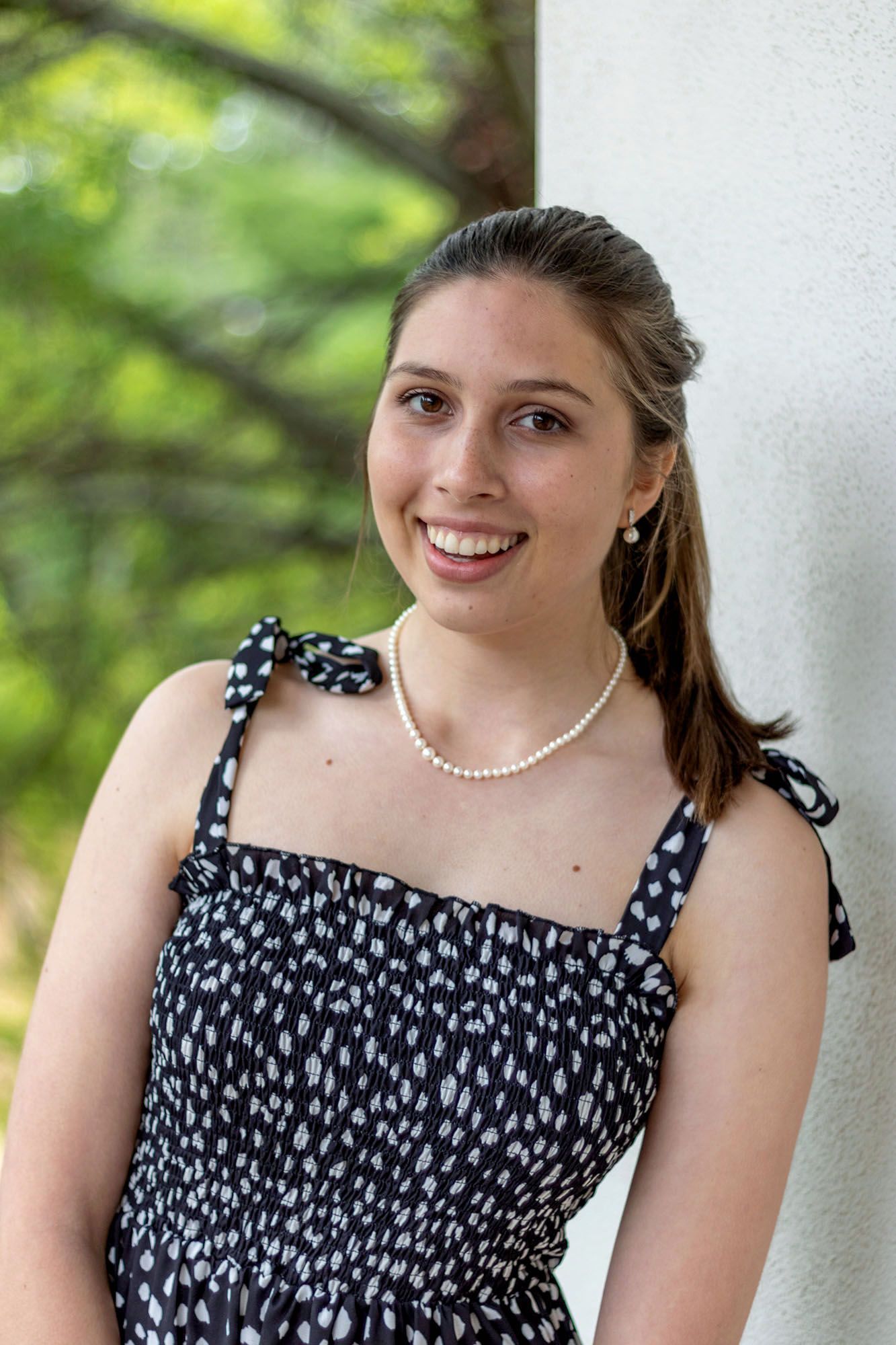 Smiling young woman in black floral sundress with pearl necklace posing outdoors near column.