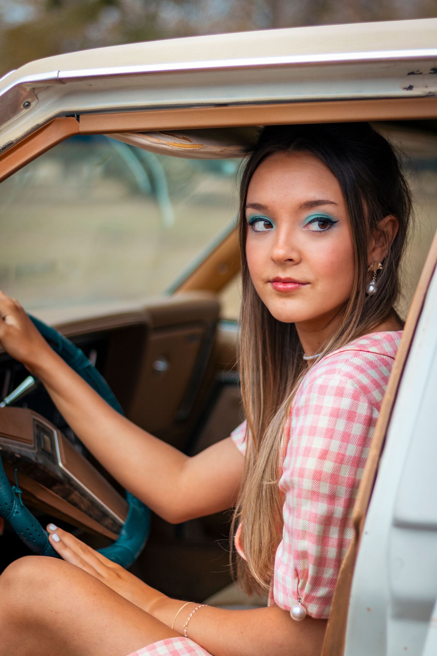 Young woman in pink gingham dress with teal eye makeup sits in vintage car interior.